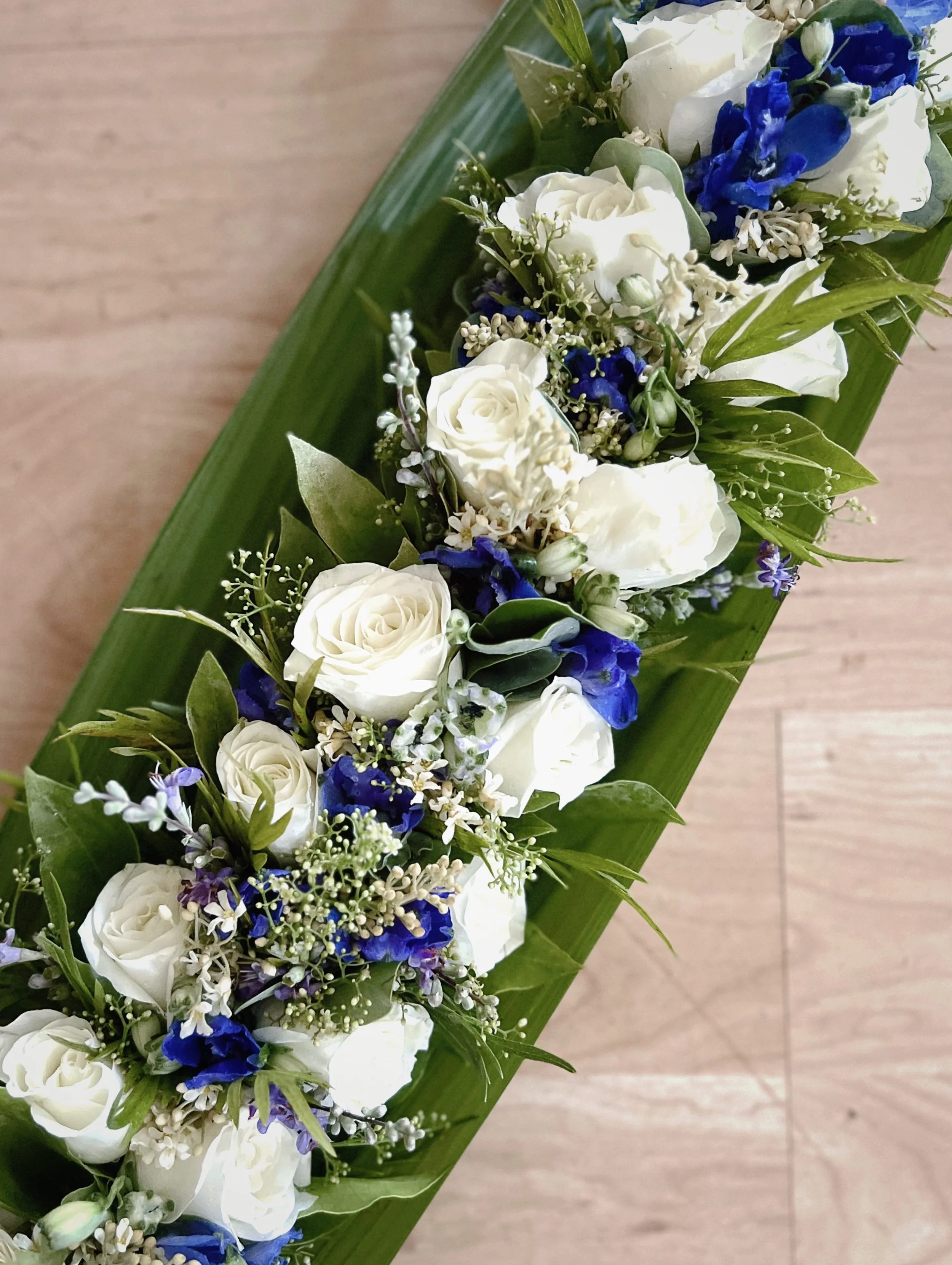 Floral arrangement with white roses, blue flowers, greenery, and small white filler flowers on a wooden background.