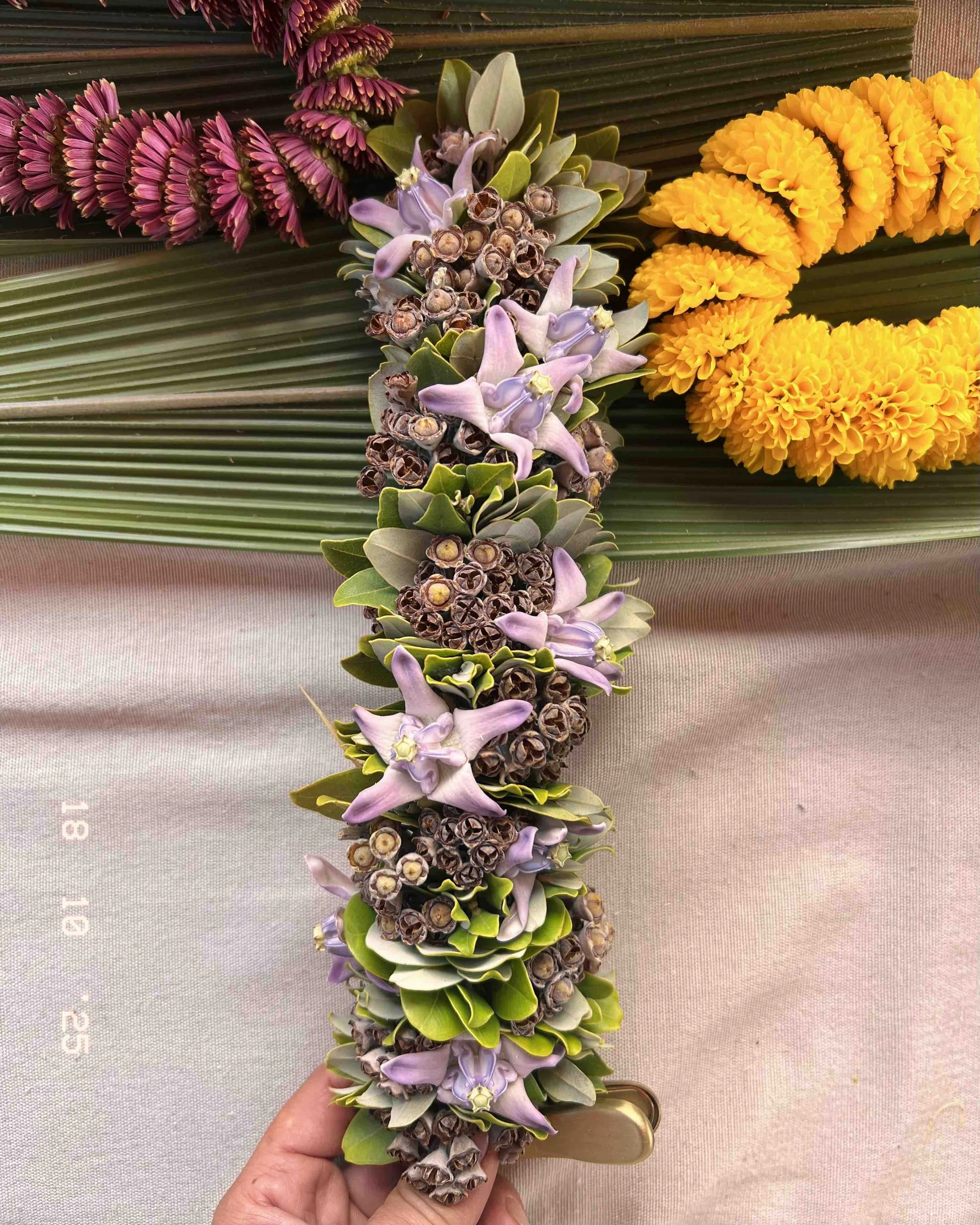 A floral garland with purple, green, and brown flowers, being held by a person's hand, with yellow marigold flowers and palm leaves in the background.