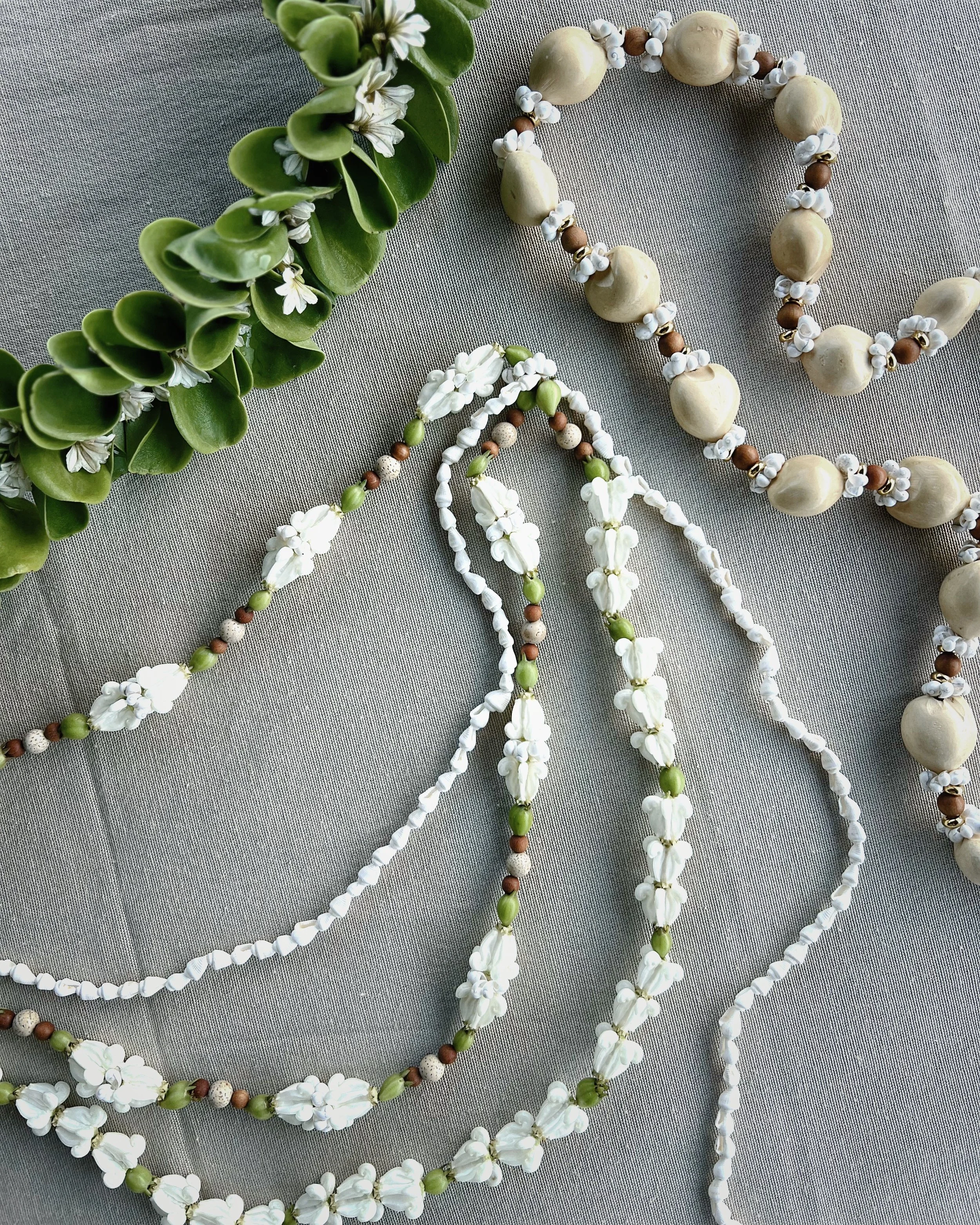 Multiple beaded necklaces in shades of white, green, and brown laid on a gray fabric surface, with a green leafy plant with white flowers in the top left corner.