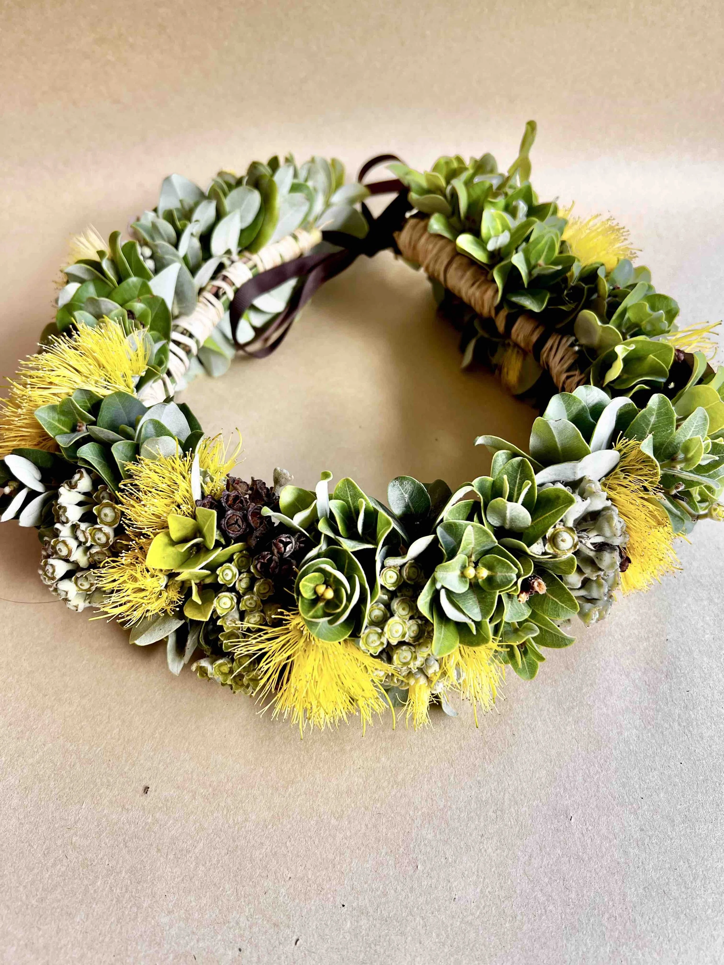 A floral wreath made with green leaves, yellow flowers, and white berries, decorated with a dark ribbon.