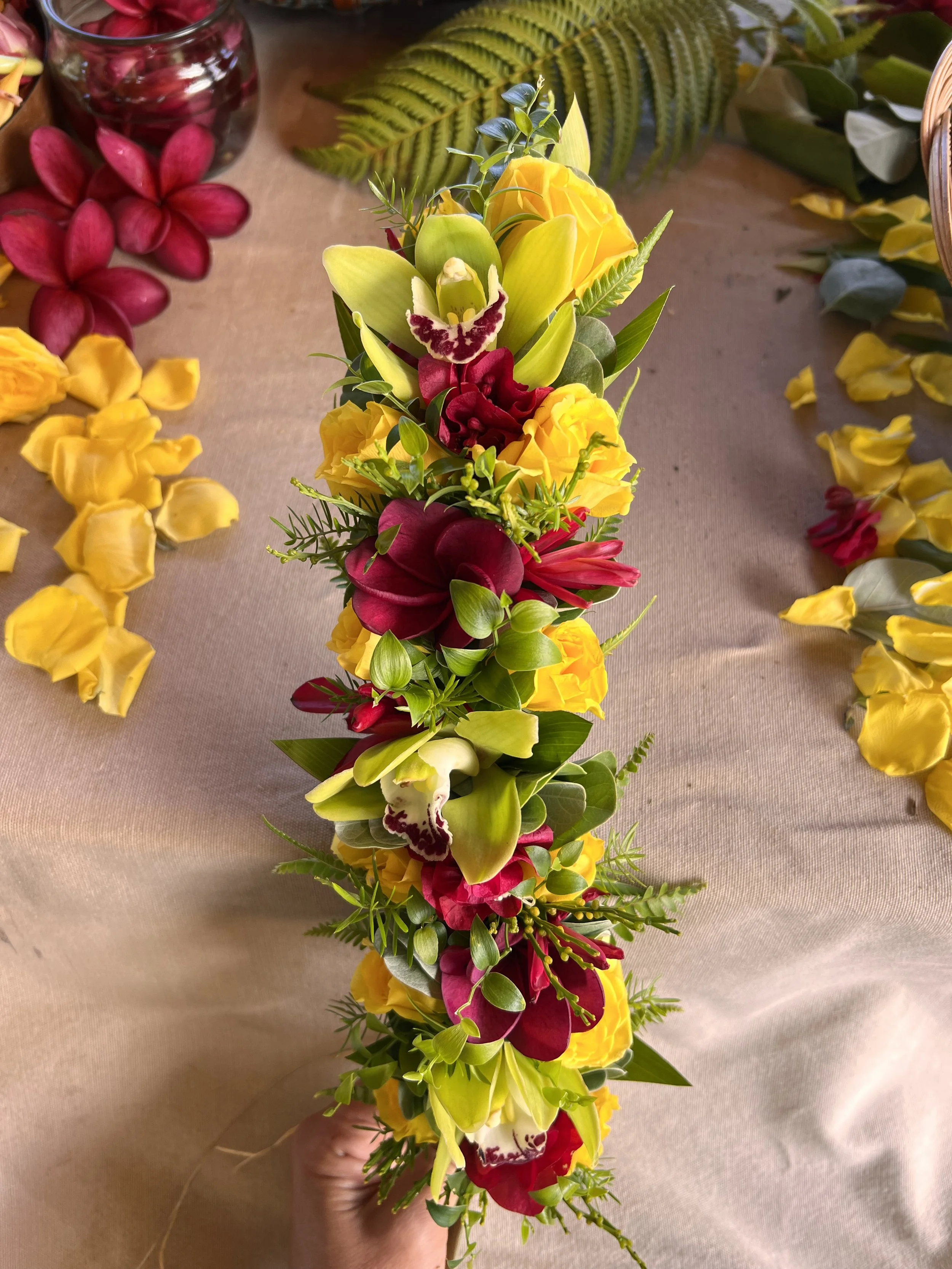 A vibrant floral arrangement with yellow, red, and white orchids, surrounded by green foliage, held by a hand over a beige surface, with additional scattered flowers visible around.