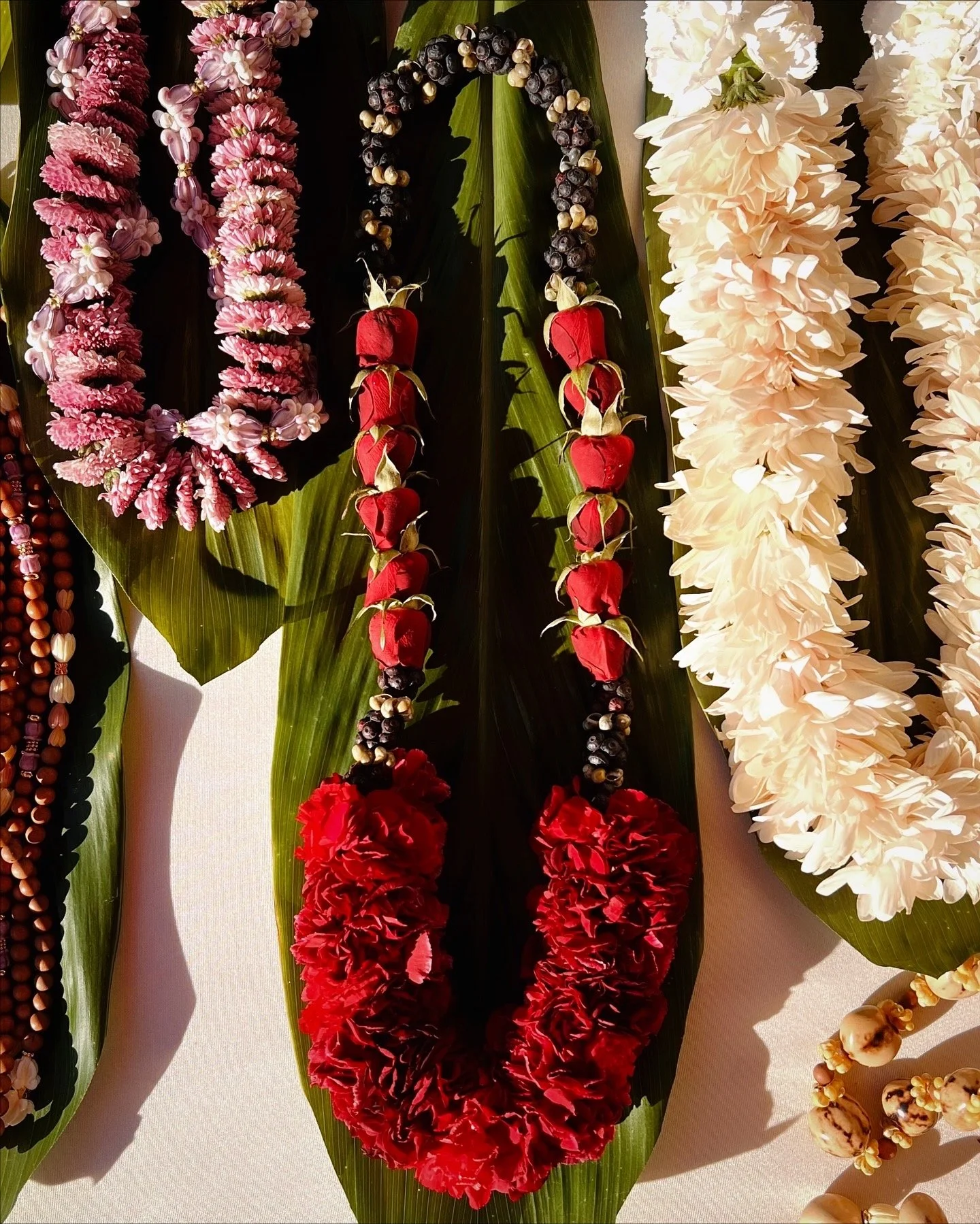 Multiple floral leis and necklaces made of flowers, berries, and seeds displayed on green leaves.