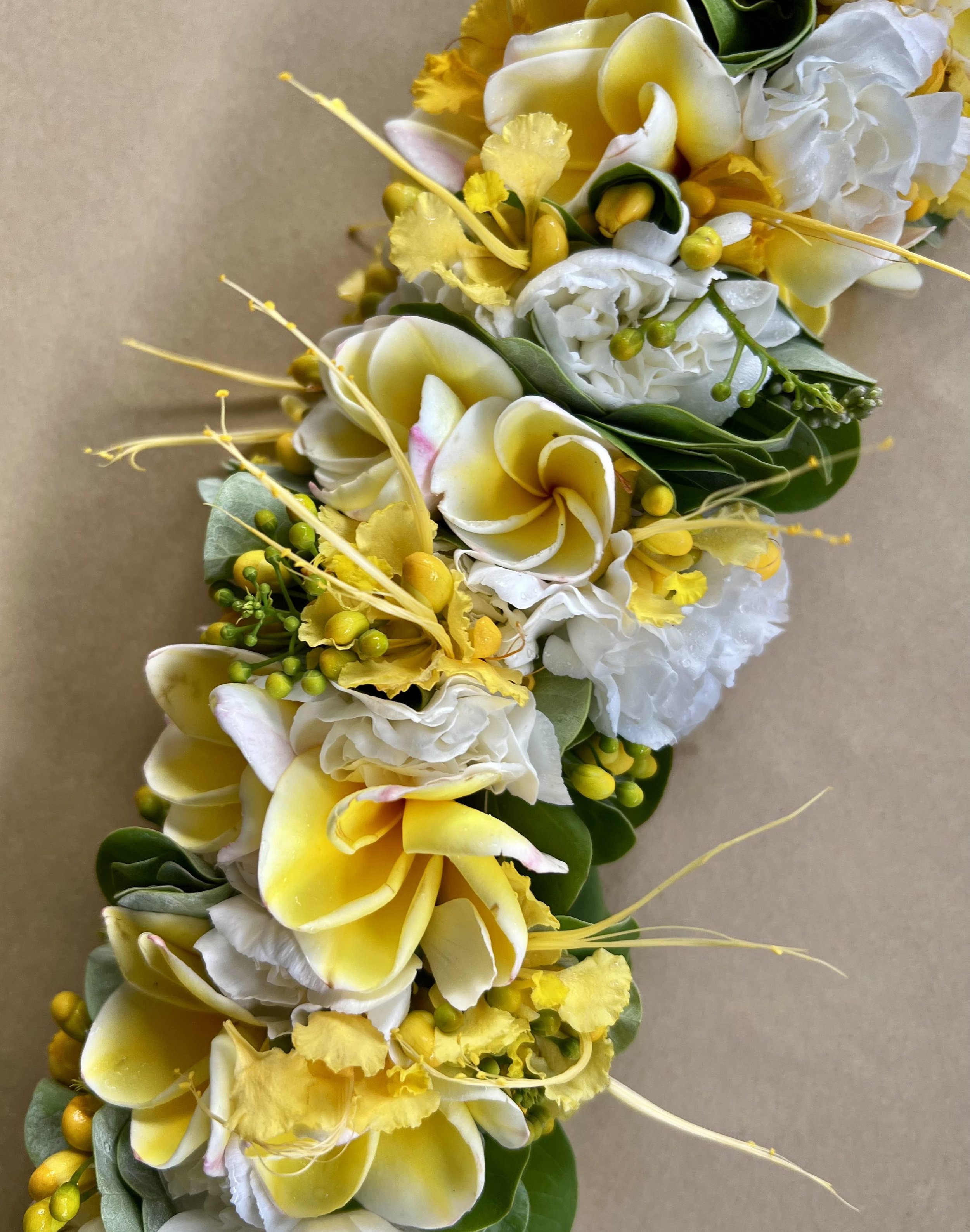 A close-up of a floral arrangement consisting of yellow and white flowers, including calla lilies, roses, and small yellow buds, with green leaves on a beige background.