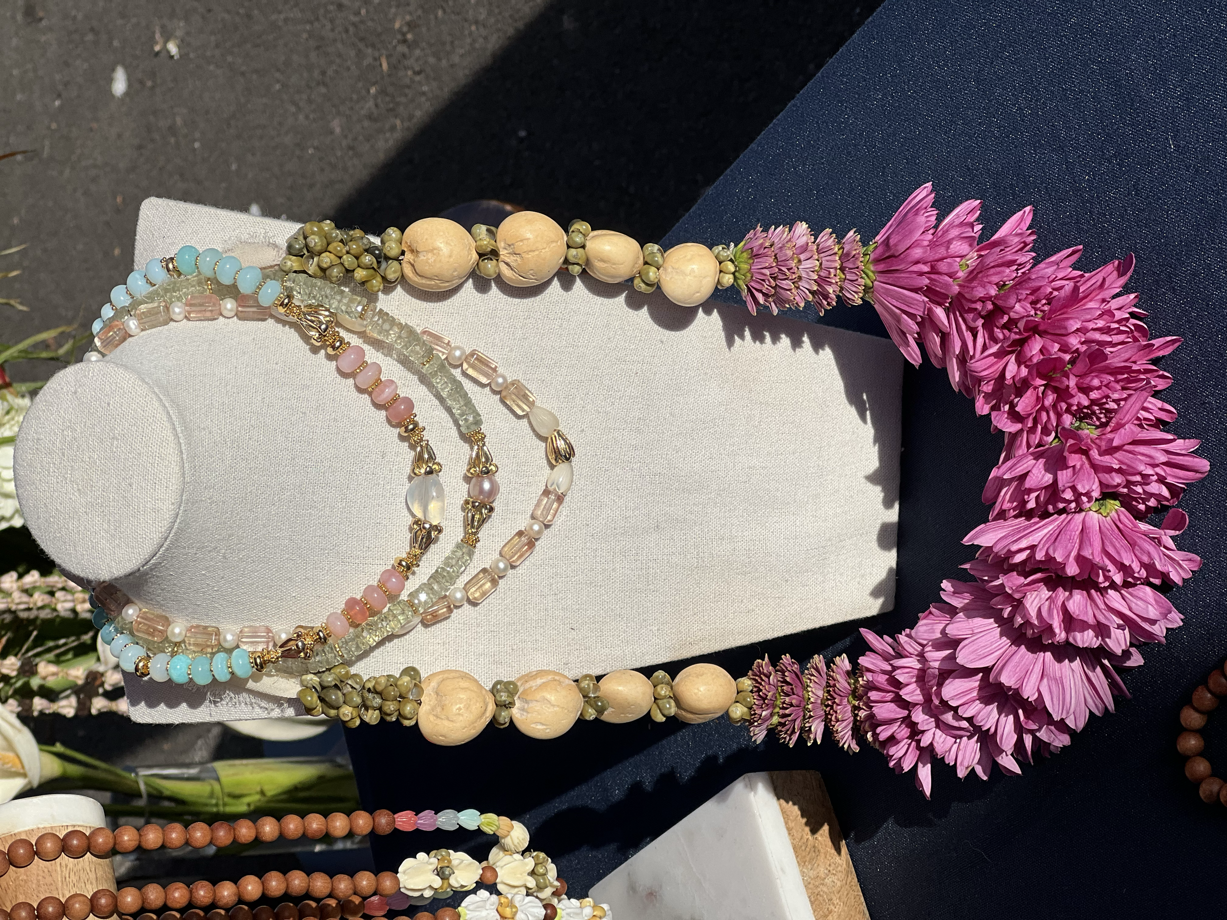 A jewelry display with a necklace of pink, white, and blue beads, and a floral necklace made of pink flowers, on a white and dark blue surface.