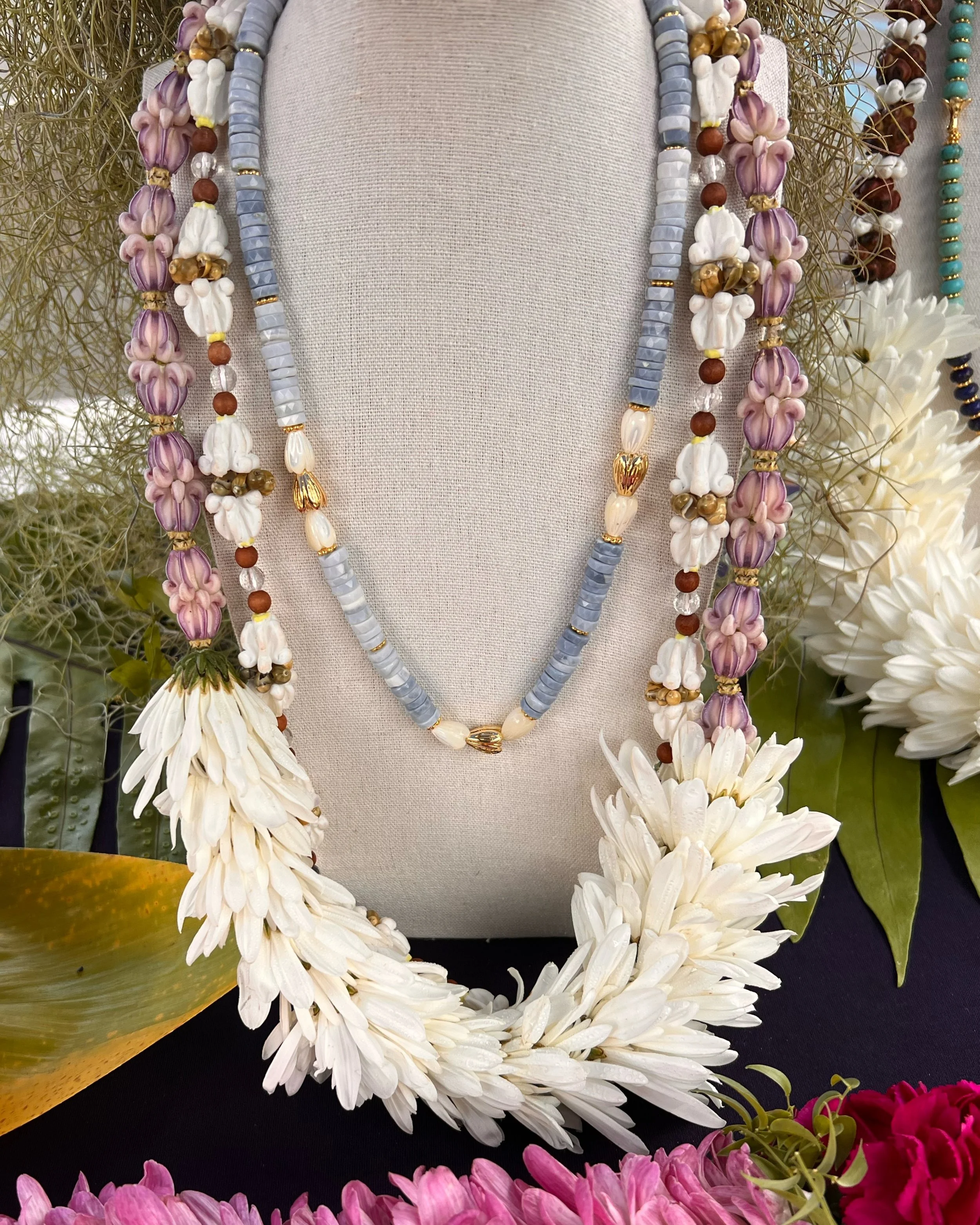 Display of various beaded necklaces arranged around a cloth-covered stand, with some necklaces featuring purple, white, blue, and gold beads, surrounded by white and pink flowers and greenery.