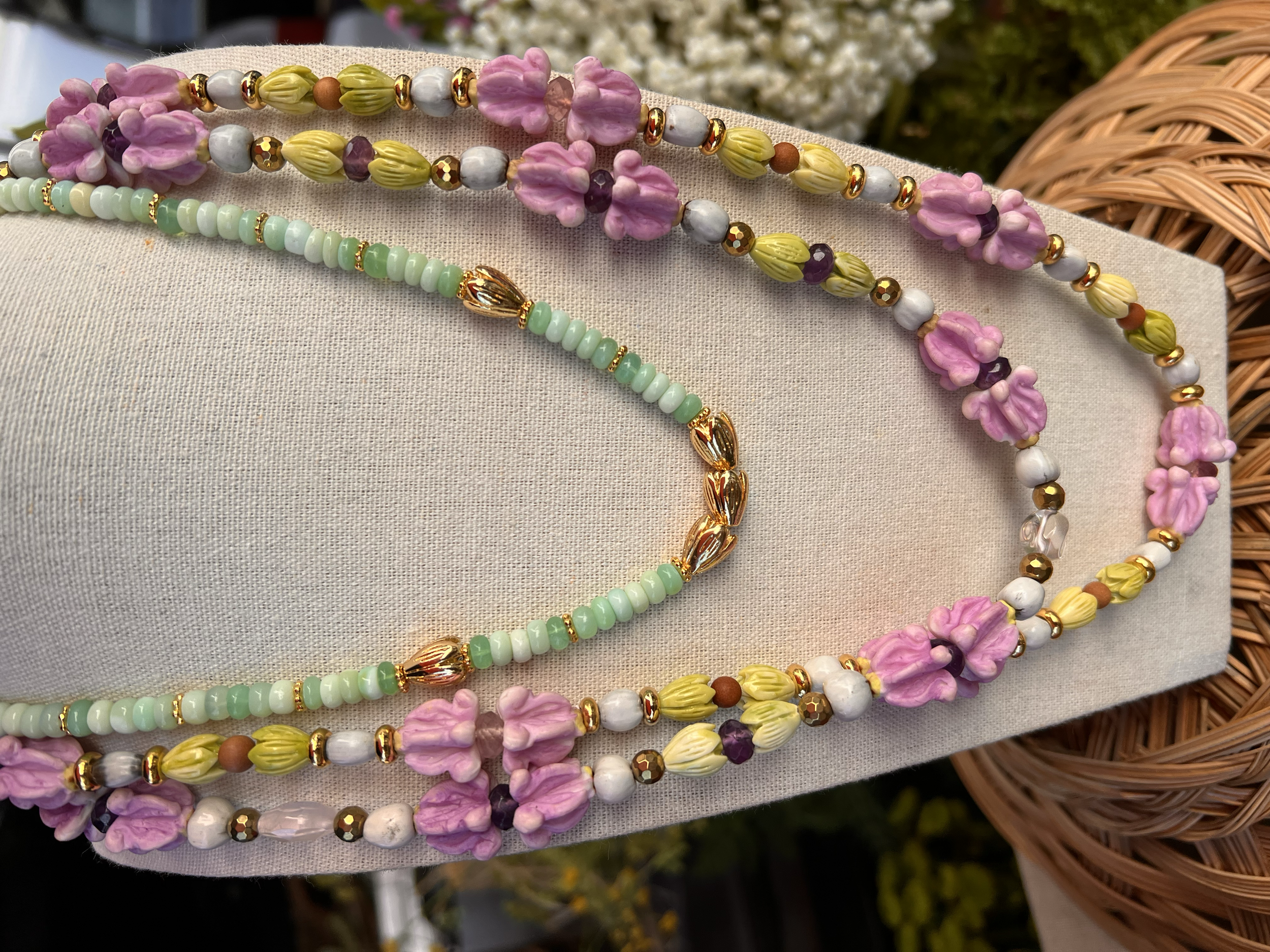 Two beaded necklaces displayed on a beige fabric stand, one with pink flower-shaped beads, yellow and green beads, and gold accents, the other with small light green beads, gold accents, and larger gold beads, against a backdrop of wicker basket and 