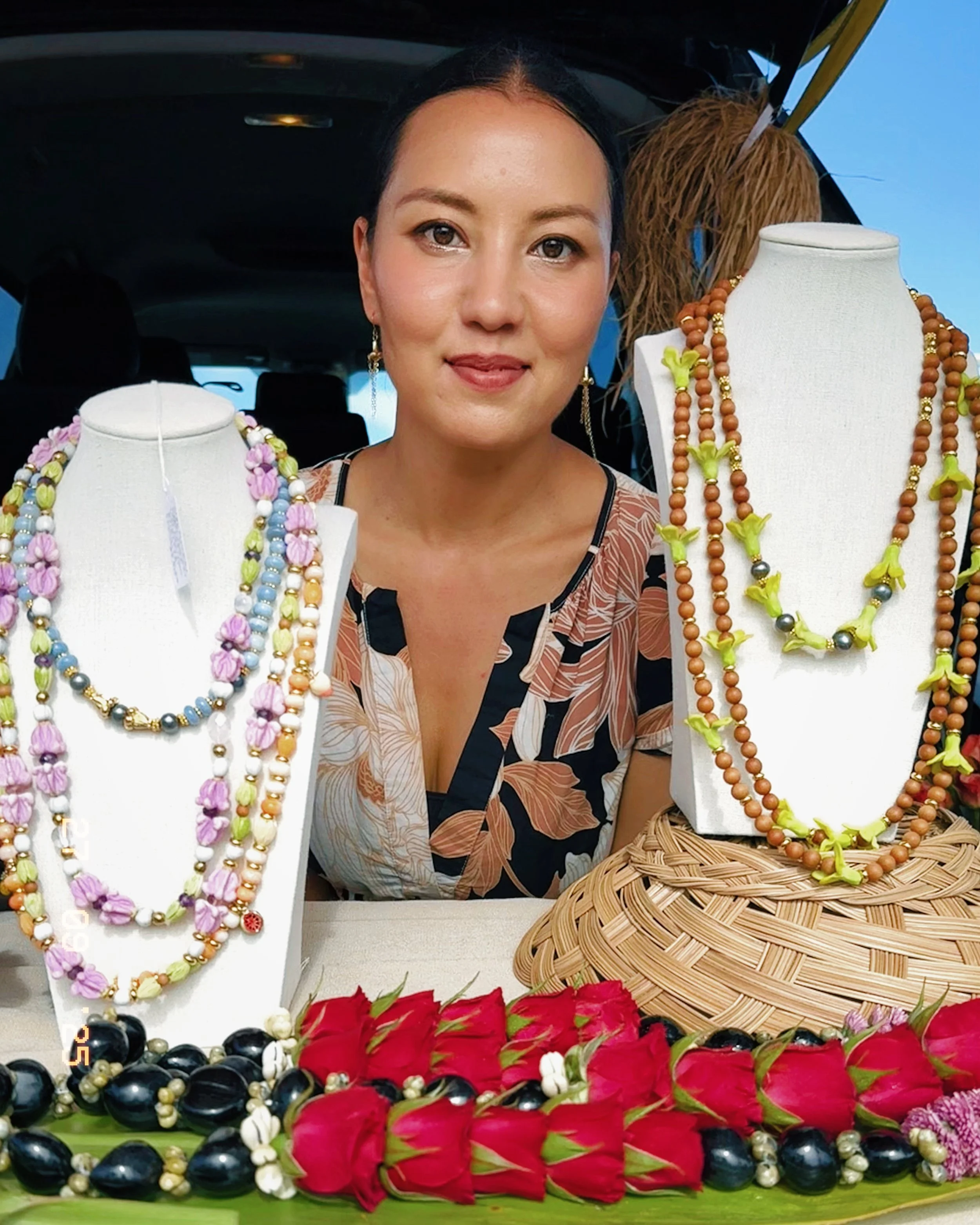 A woman sitting at a jewelry display with necklaces on white busts, colorful flowers and beads on the table.