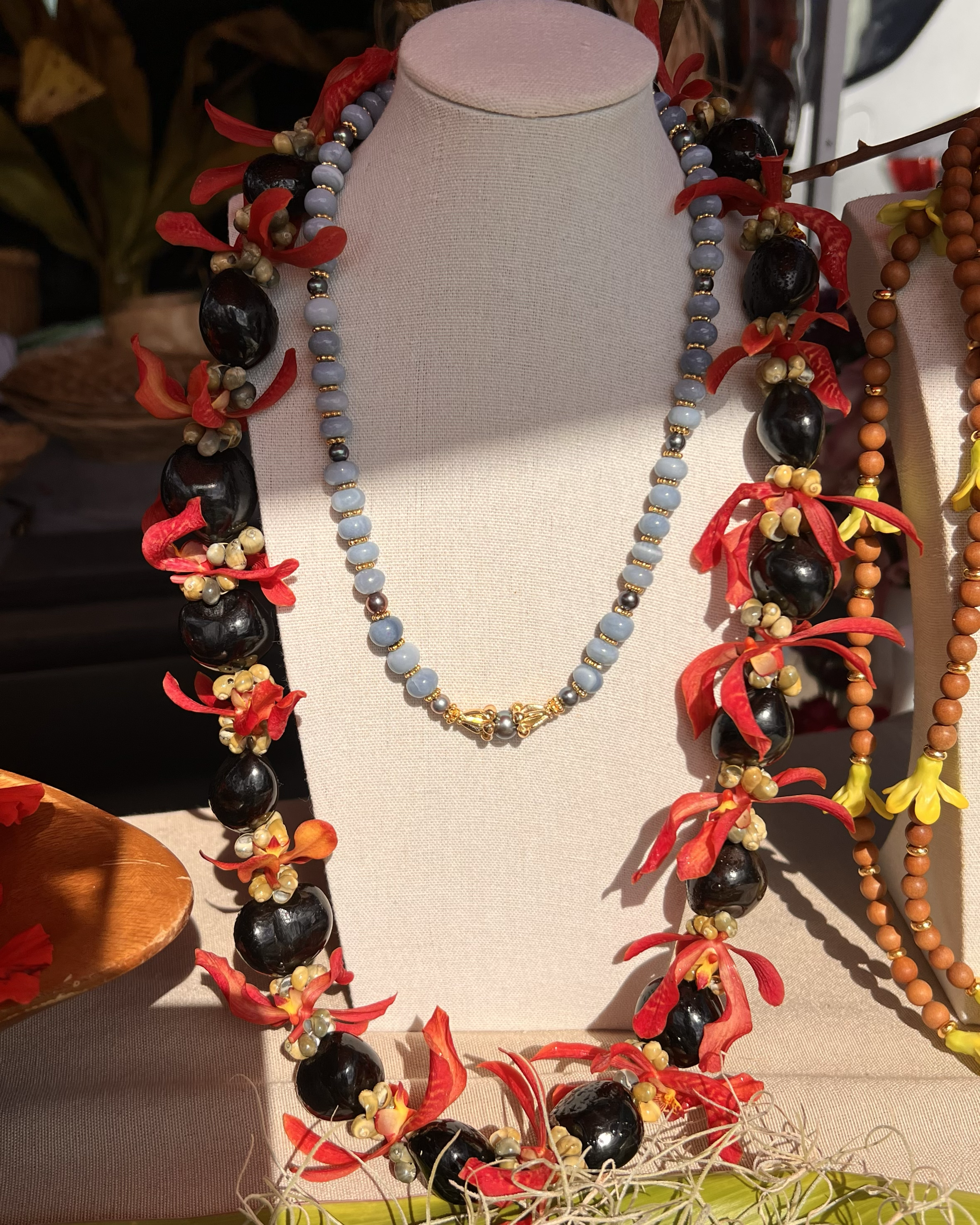 Display of various beaded necklaces on a beige bust, including a light blue beaded necklace, and a large black beaded necklace with red and yellow floral accents.