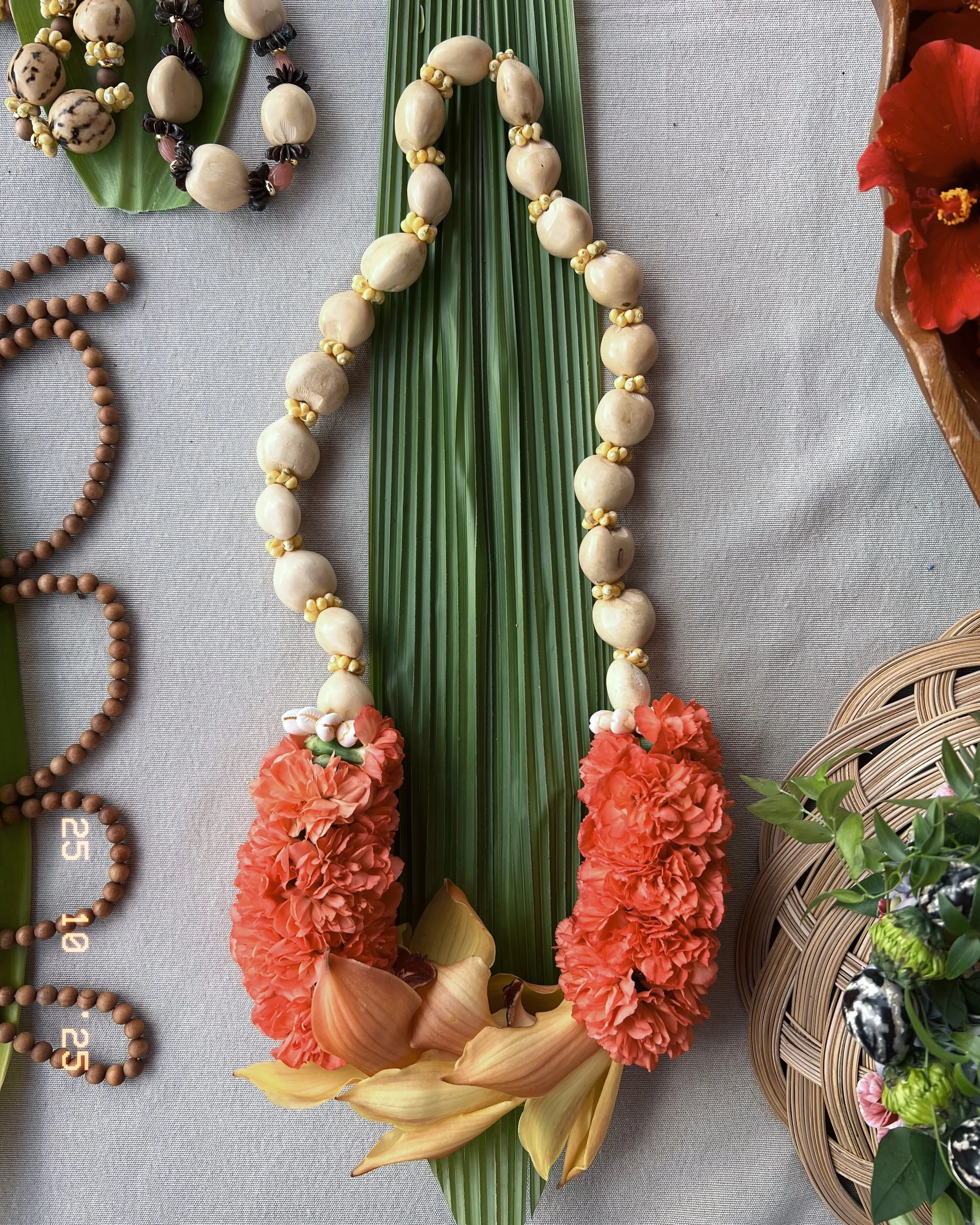 Traditional floral garland with large orange carnations, smaller orange and white flowers, and ivory beads, laid over a green leaf on a gray fabric surface.