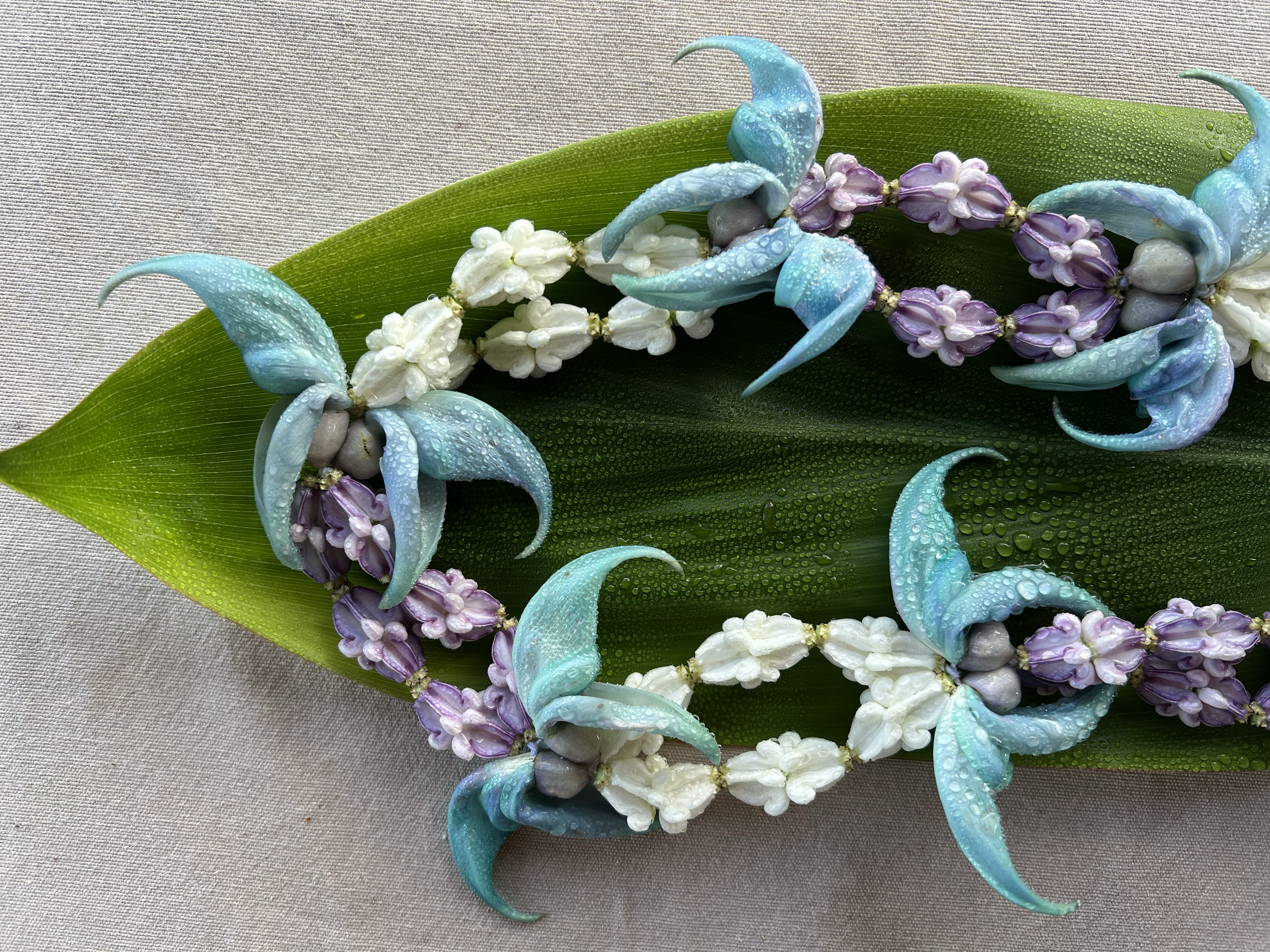 A flower necklace with blue, purple, and white flowers, placed on a large green leaf with water droplets on it.