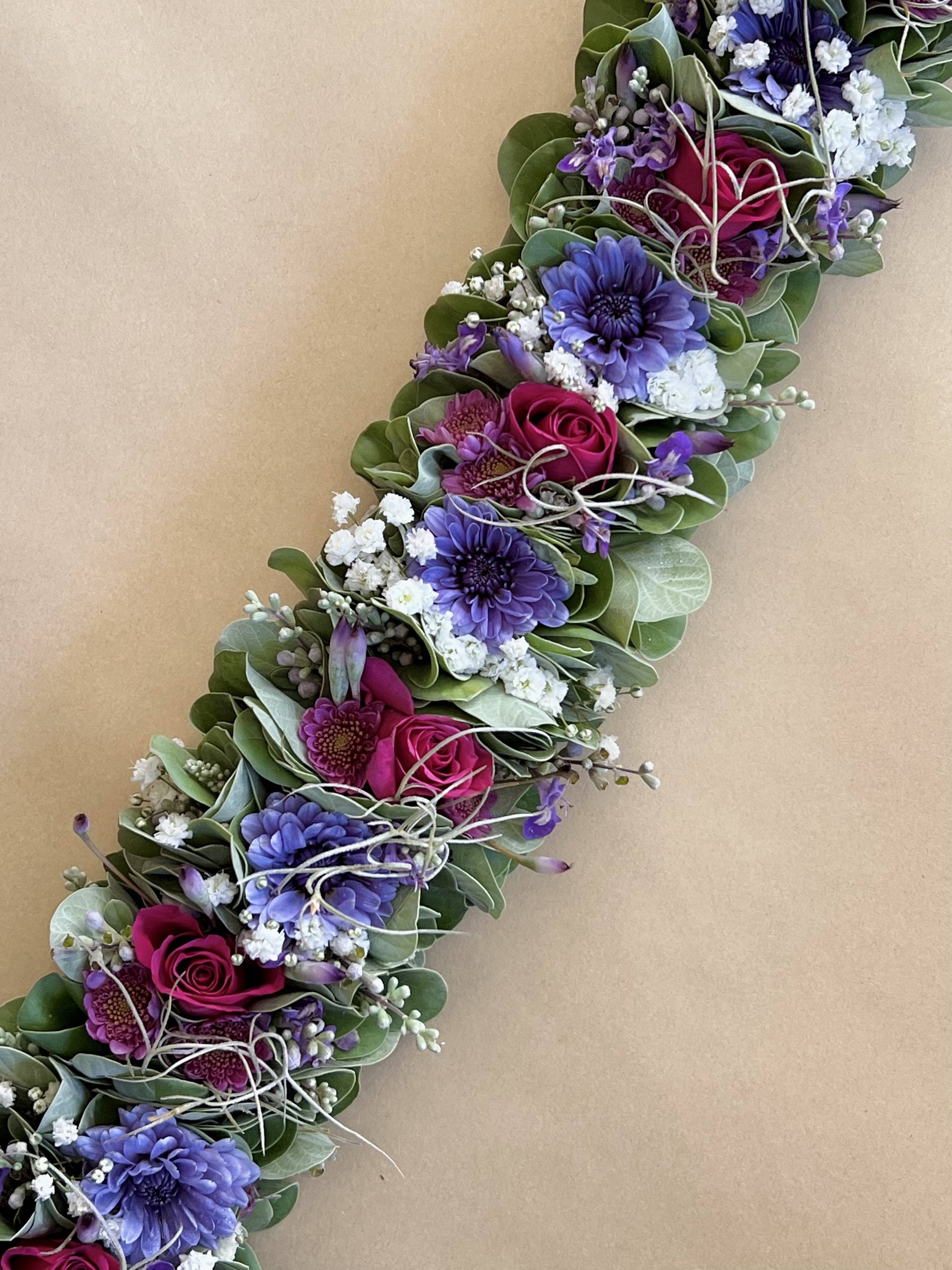 A floral arrangement with pink roses, purple dahlias, white baby's breath, and green leaves arranged in a diagonal line on a beige background.