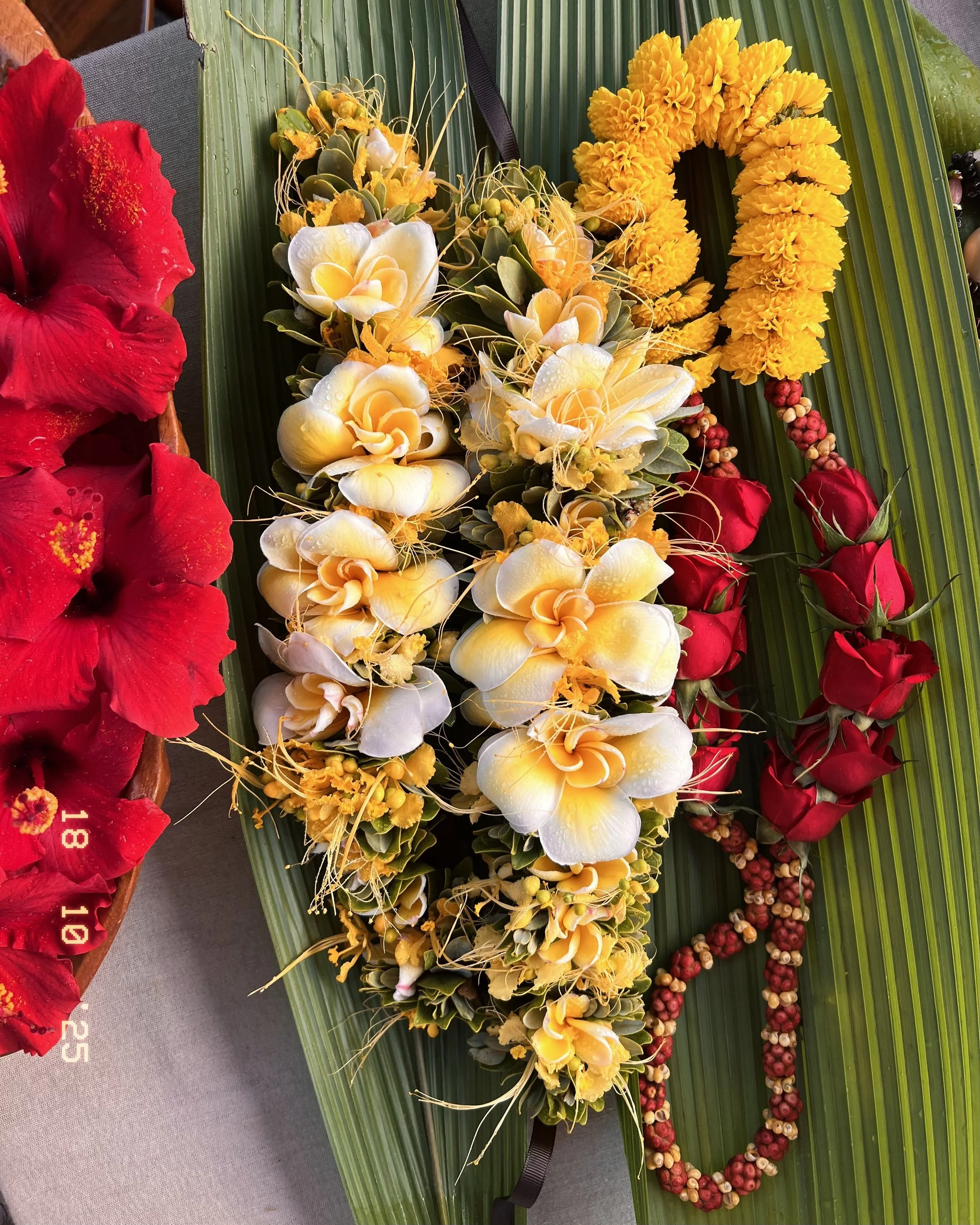 Hawaiian floral lei with yellow and white flowers, red roses, and greenery, displayed on a large green palm leaf.