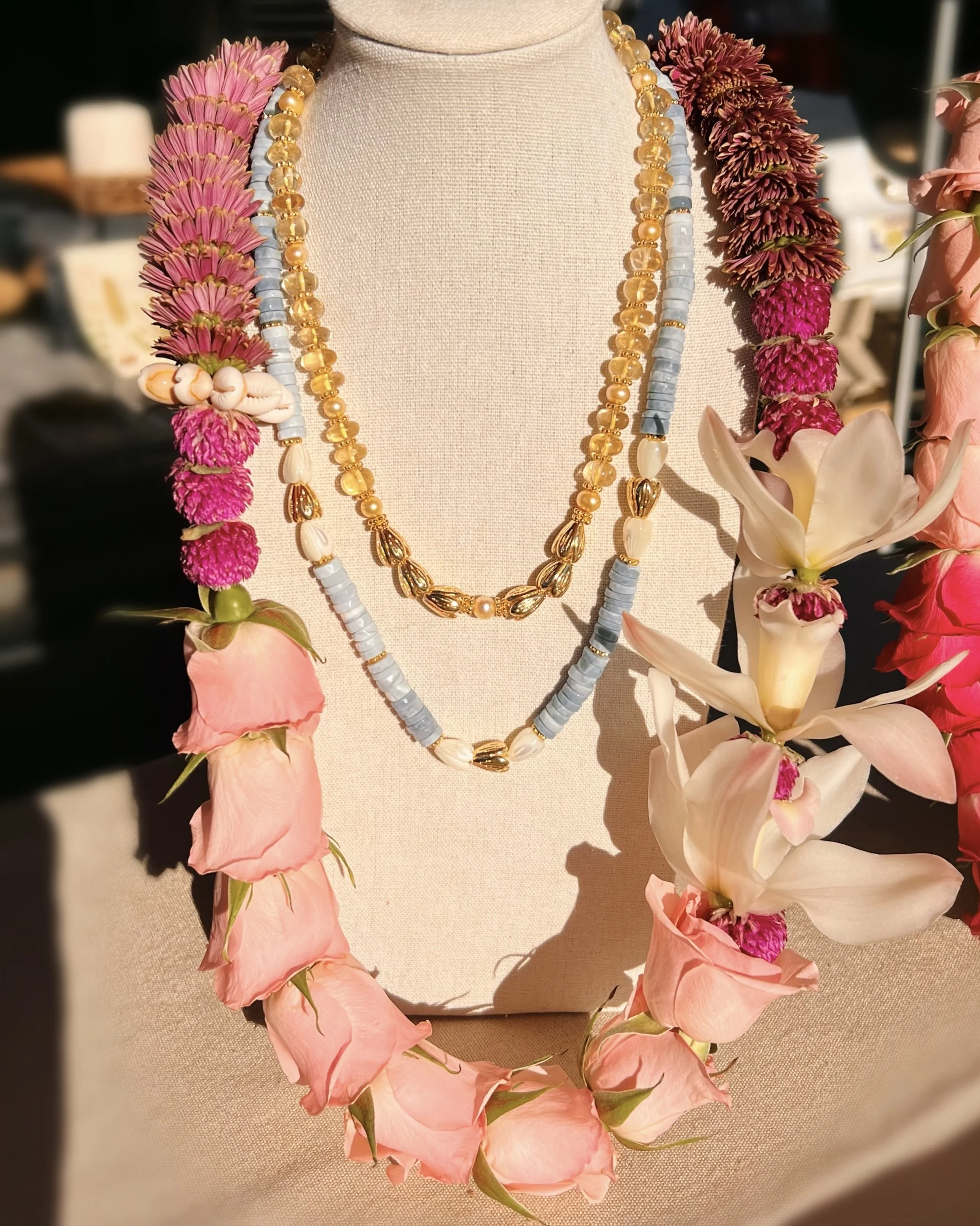 Display of three layered necklaces on a jewelry bust surrounded by pink, purple, and white flower leis.