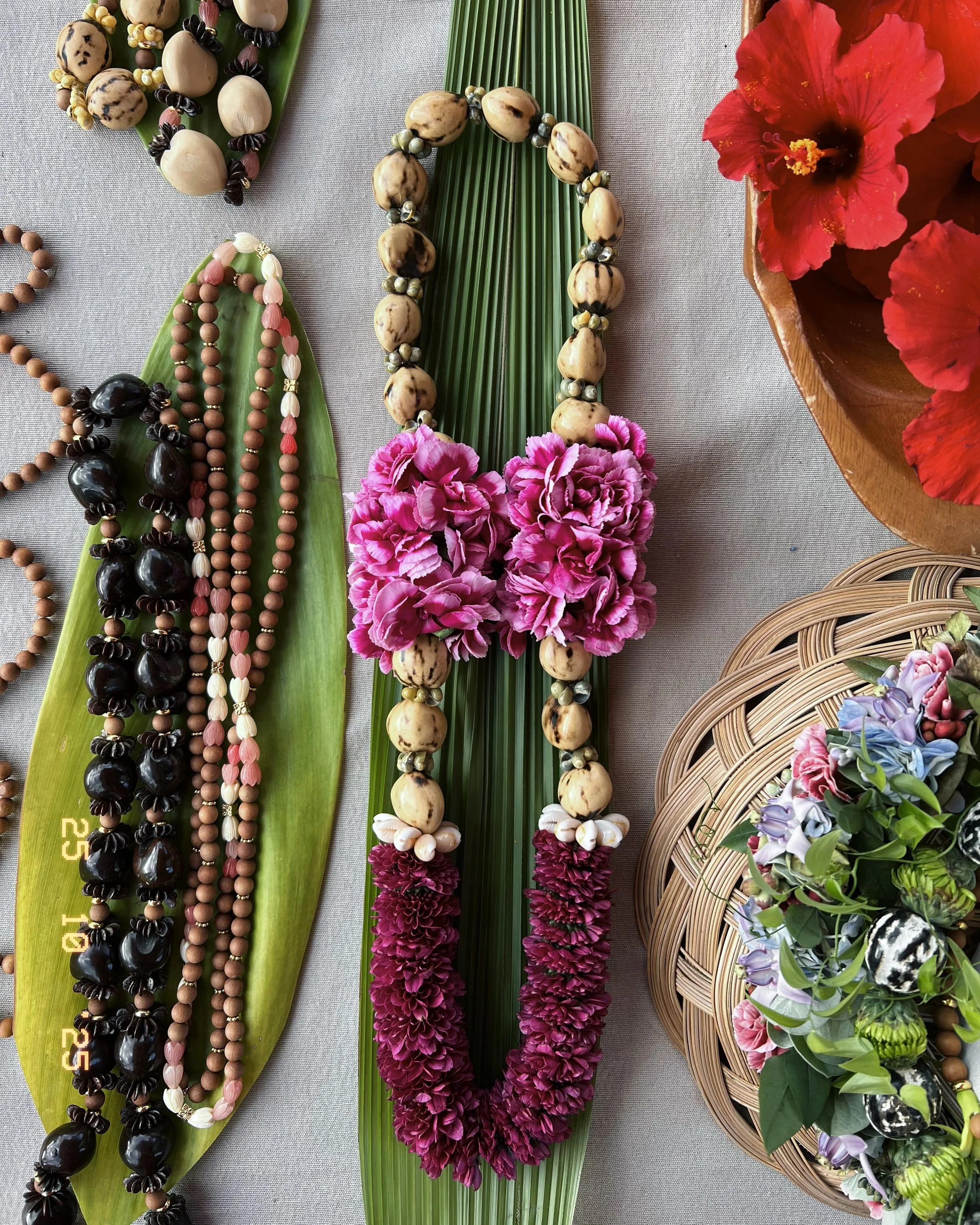 Colorful floral necklaces and leis displayed on green leaves and a gray surface, with red and purple flowers in background.