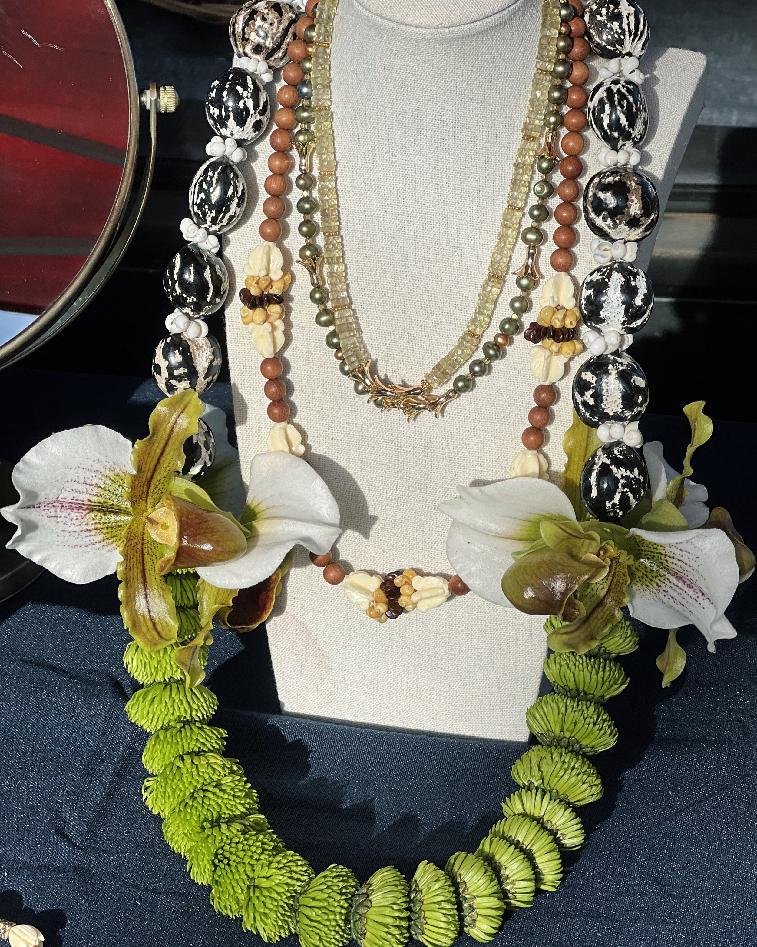Display of three layered necklaces on a stand, featuring beads and floral accents, with a large green floral necklace and orchids at the base.