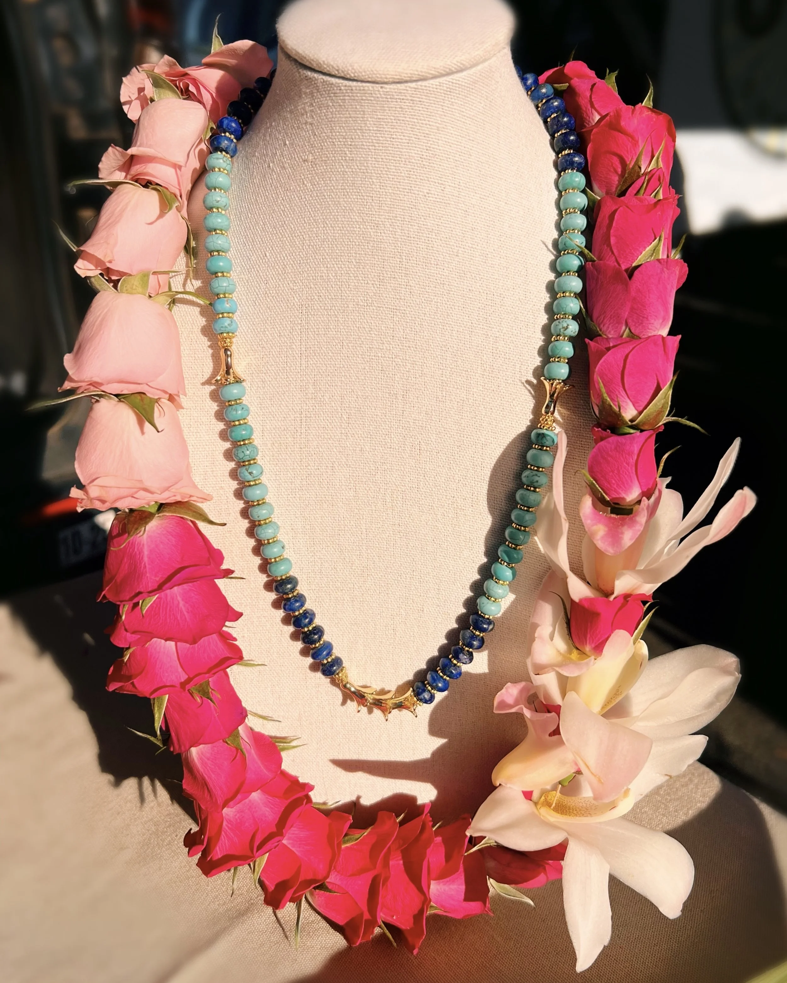 Turquoise and dark blue beaded necklace displayed on a beige bust, surrounded by pink and white flower leis with green leaves.