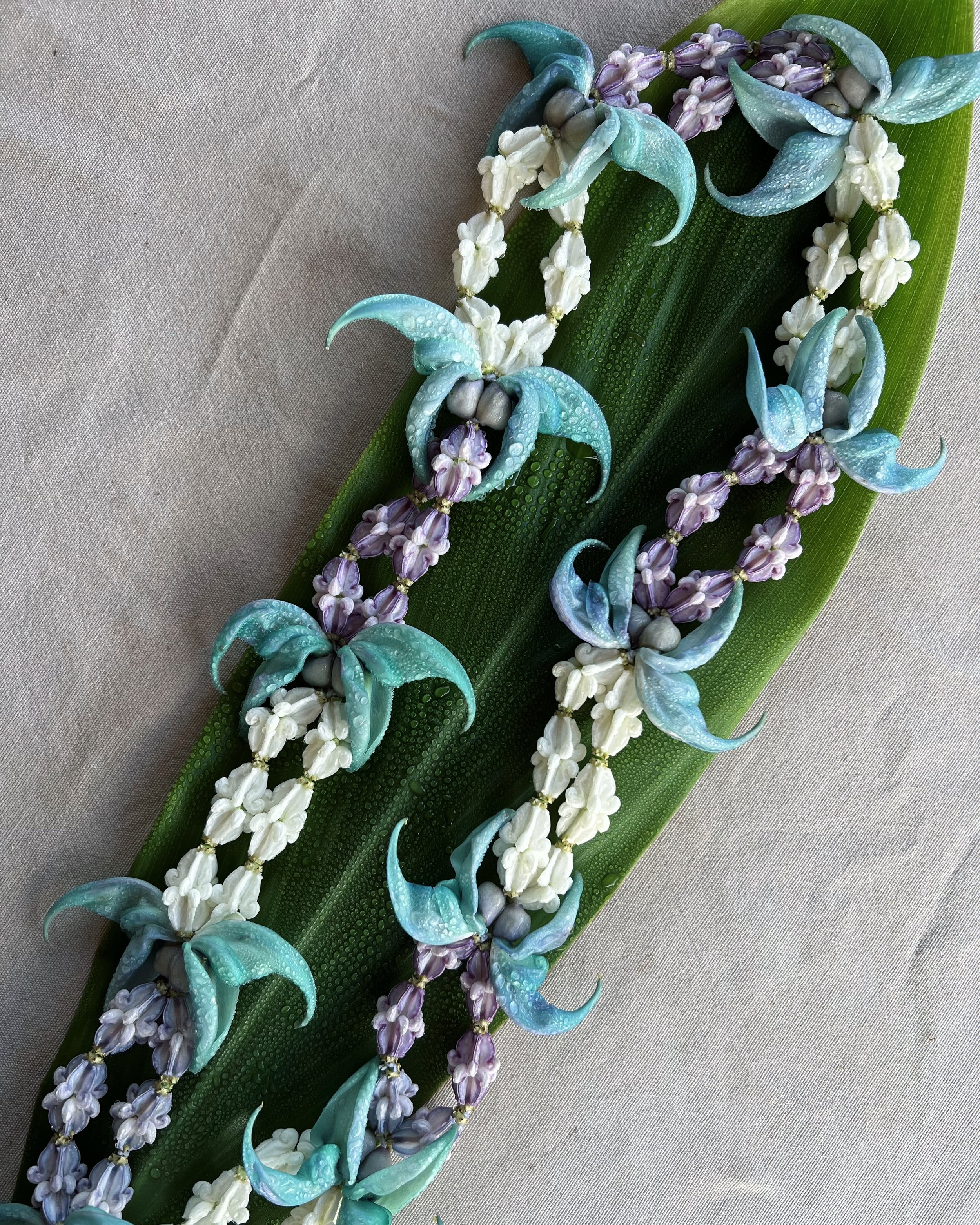 Colorful floral necklace with blue, purple, and white flowers on a green leaf resting on a beige surface.