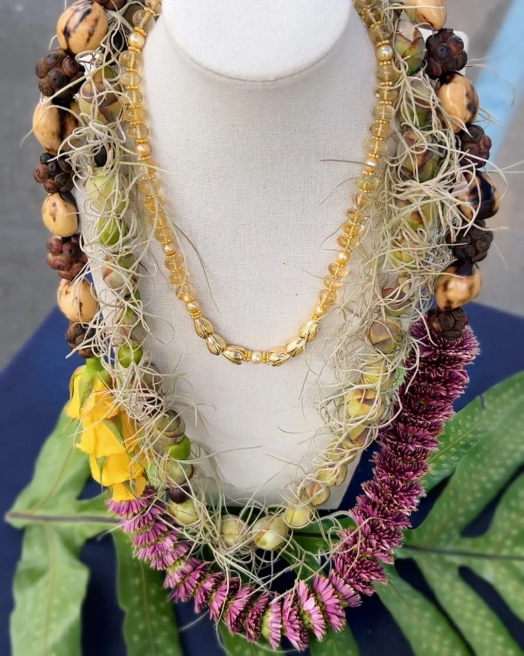 A display of multiple layered necklaces on a mannequin bust. The necklaces feature gold beads and pearls with tan and cream-colored accents. They are surrounded by floral leis made of pink and purple flowers, green leaves, and other natural materials