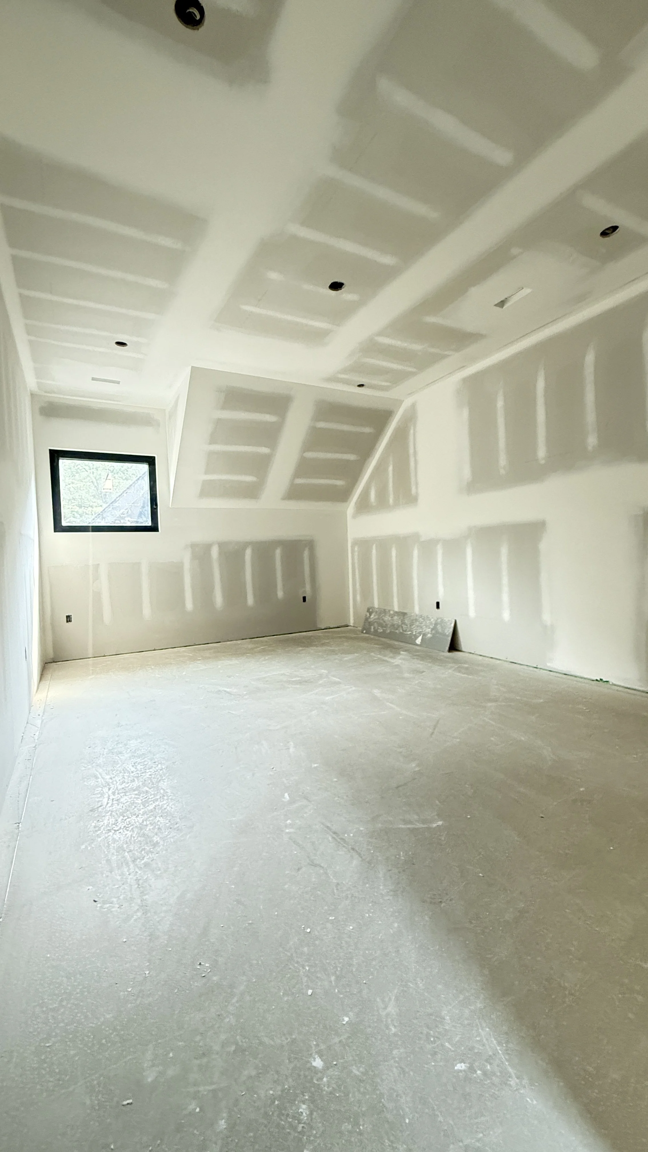 An unfinished room with drywall installed on the walls and ceiling, and a window on the left wall. The floor is bare concrete.