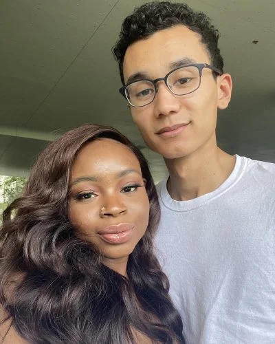 Mope and Robert taking a selfie together outside, standing under a concrete structure with greenery in the background.