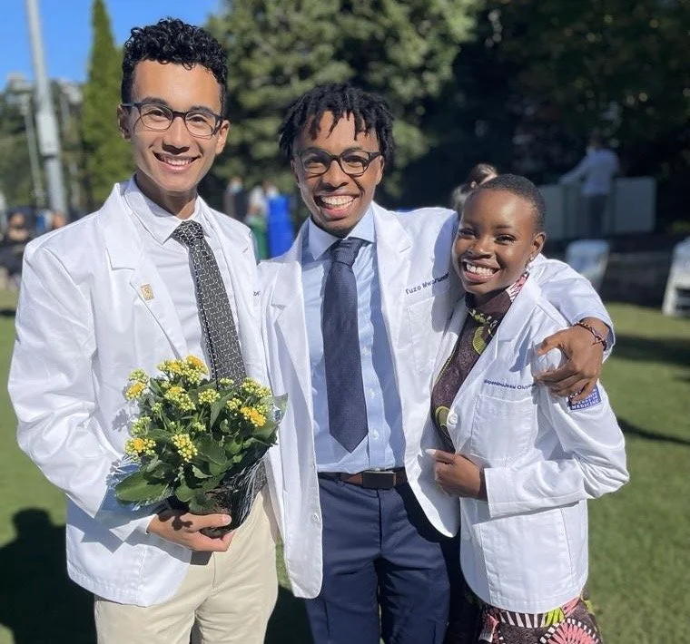 Three medical professionals outdoors smiling at the camera, two men and one woman, wearing white lab coats. The man on the left is holding a potted plant with yellow flowers, and the woman on the right has a colorful patterned dress visible under her