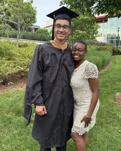 A young man in a black graduation gown and cap standing outdoors with a woman in a white lace dress, both smiling for the camera. They are on a grassy area with trees and modern buildings in the background.