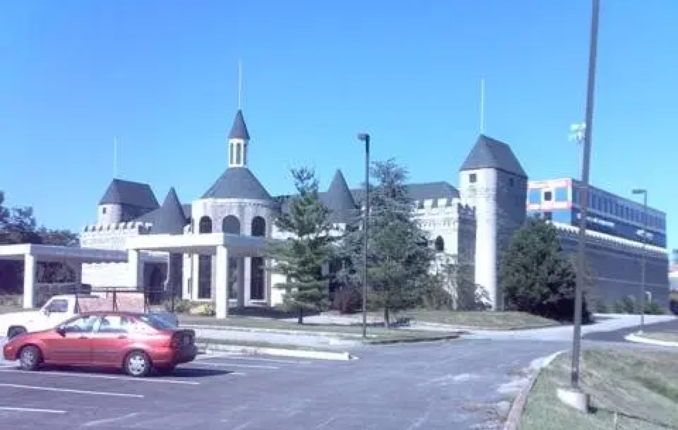 Castle-like building with turrets and towers, parking lot with cars in the foreground, trees and a clear blue sky.