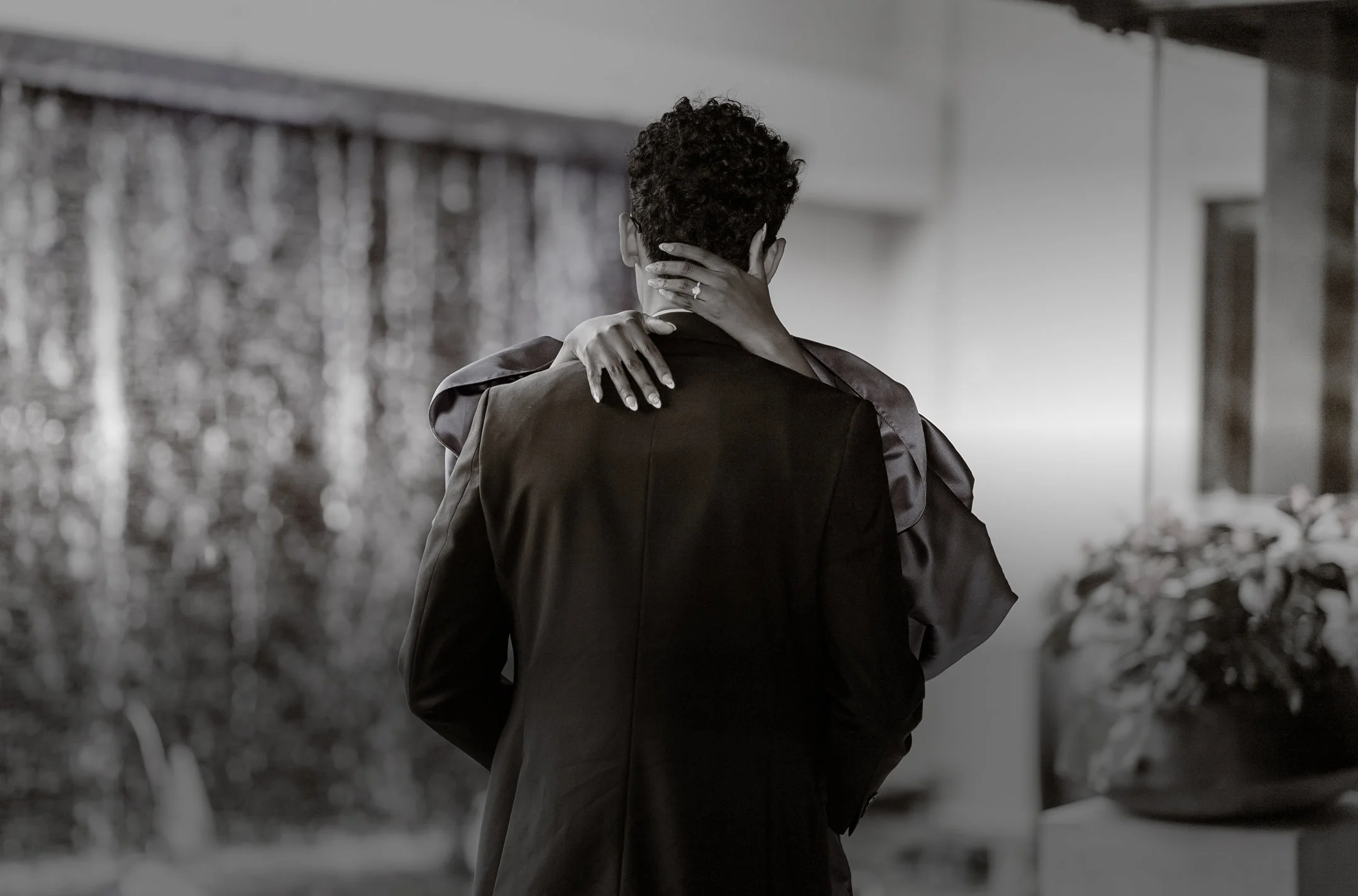 A couple embracing; Mope's arms around the Robert's shoulders, and Robert is standing with his back to the camera, indoors, with a blurred background.