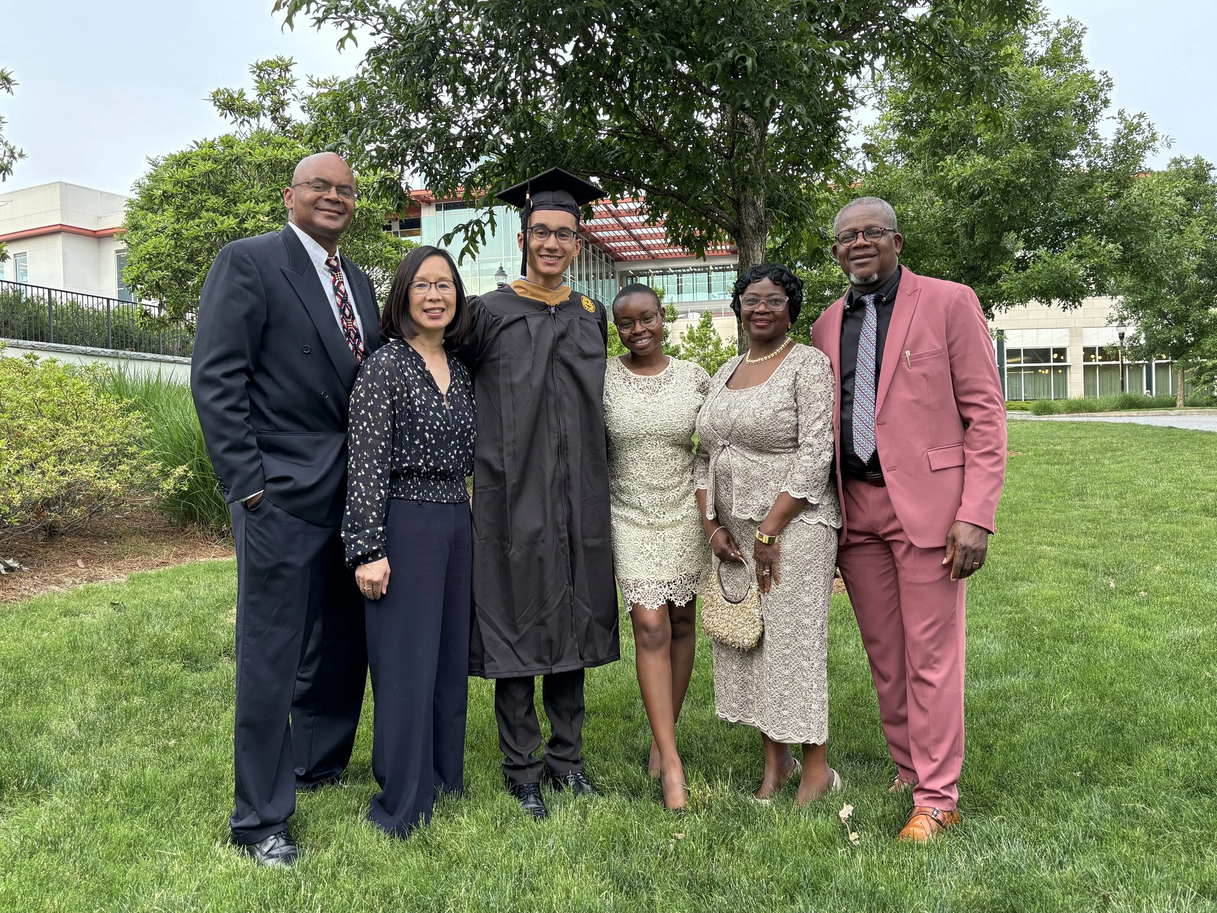 Mope and Robert standing on grass outdoors, in graduation gown and cap in the center, surrounded by Mope's parents and Robert's parents.