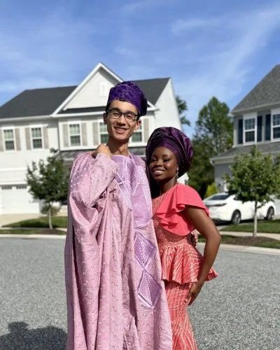 Robert and Mope dressed in traditional African attire, standing outdoors on a street with houses and cars in the background.
