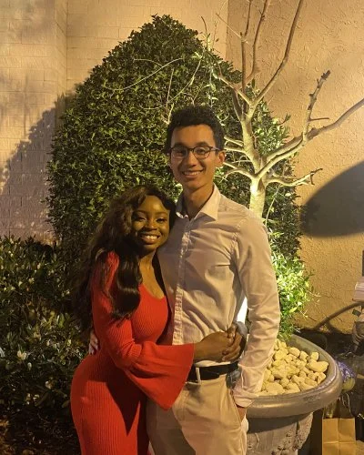 Mope and Robert posing together outdoors at night. They are standing in front of a leafy bush and a small tree, with warm lighting illuminating the scene.