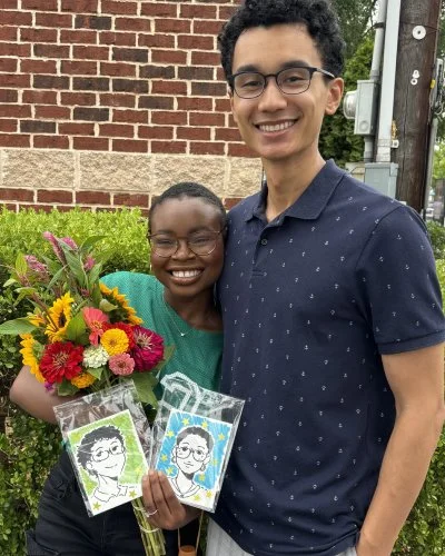 Mope holding a bouquet of colorful flowers and two cards with her illustration, standing next to Robert, both smiling outdoors in front of a brick wall and greenery.