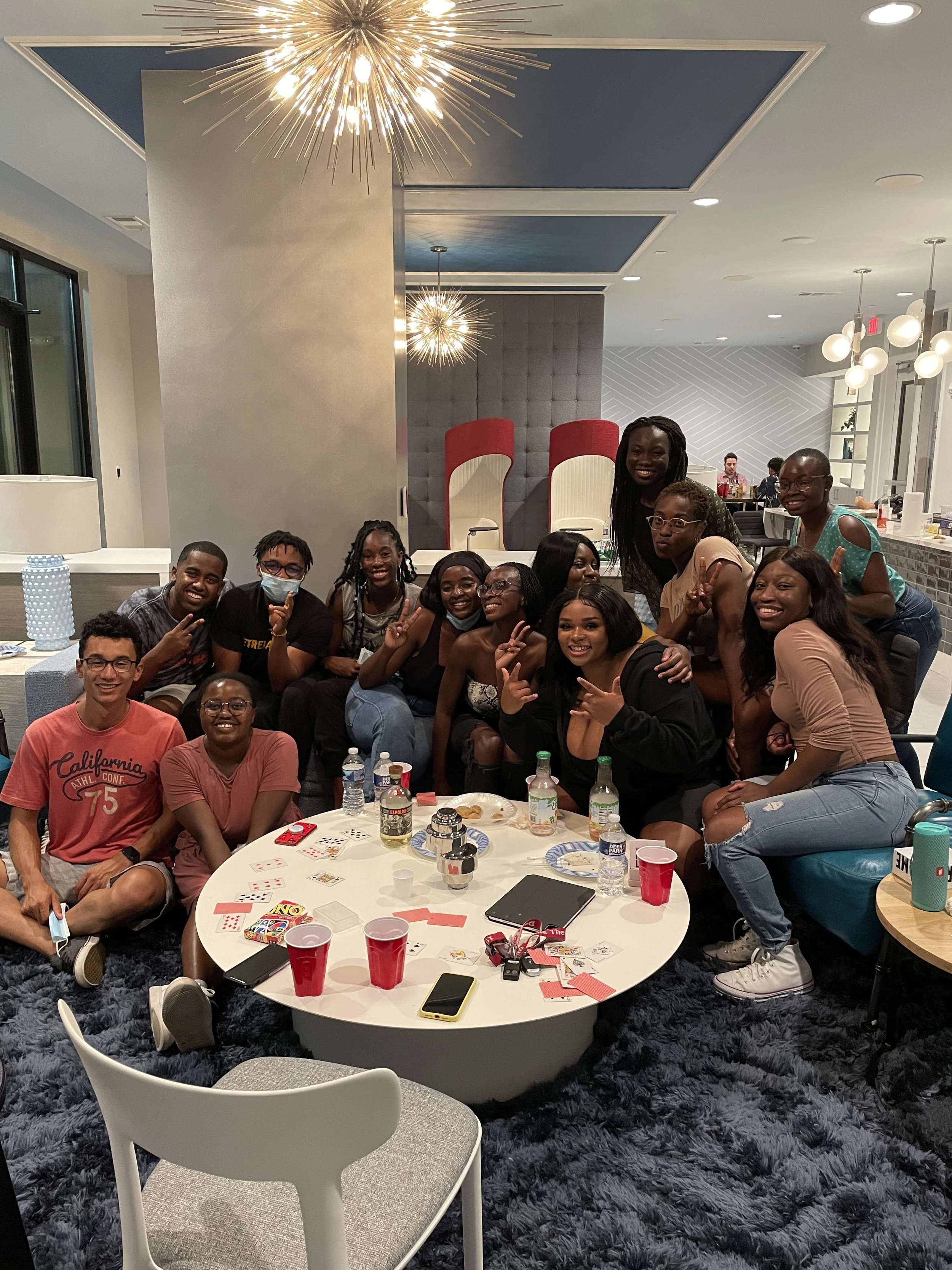 A group of Robert and Mope's friends gathered around a table in a modern indoor lounge, celebrating and smiling, with various drinks, cards, and party items on the table.