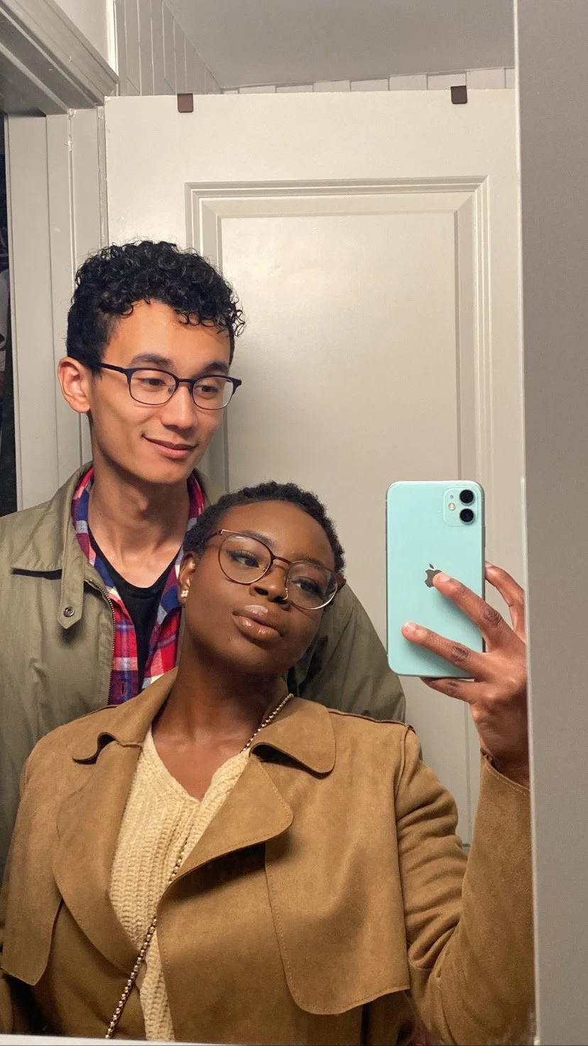 Mope taking a mirror selfie with Robert standing behind her in a room with white walls and a door.