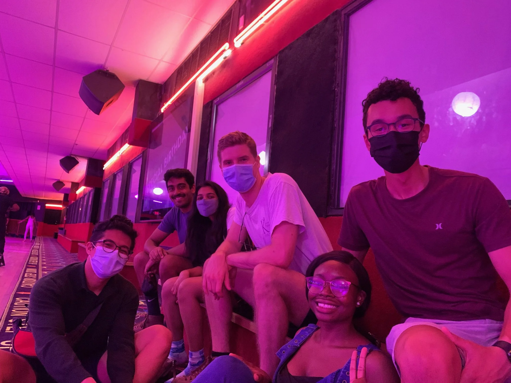Mope and Robert sitting and kneeling on the floor at an indoor roller skating rink with their friends, with purple and pink lighting.