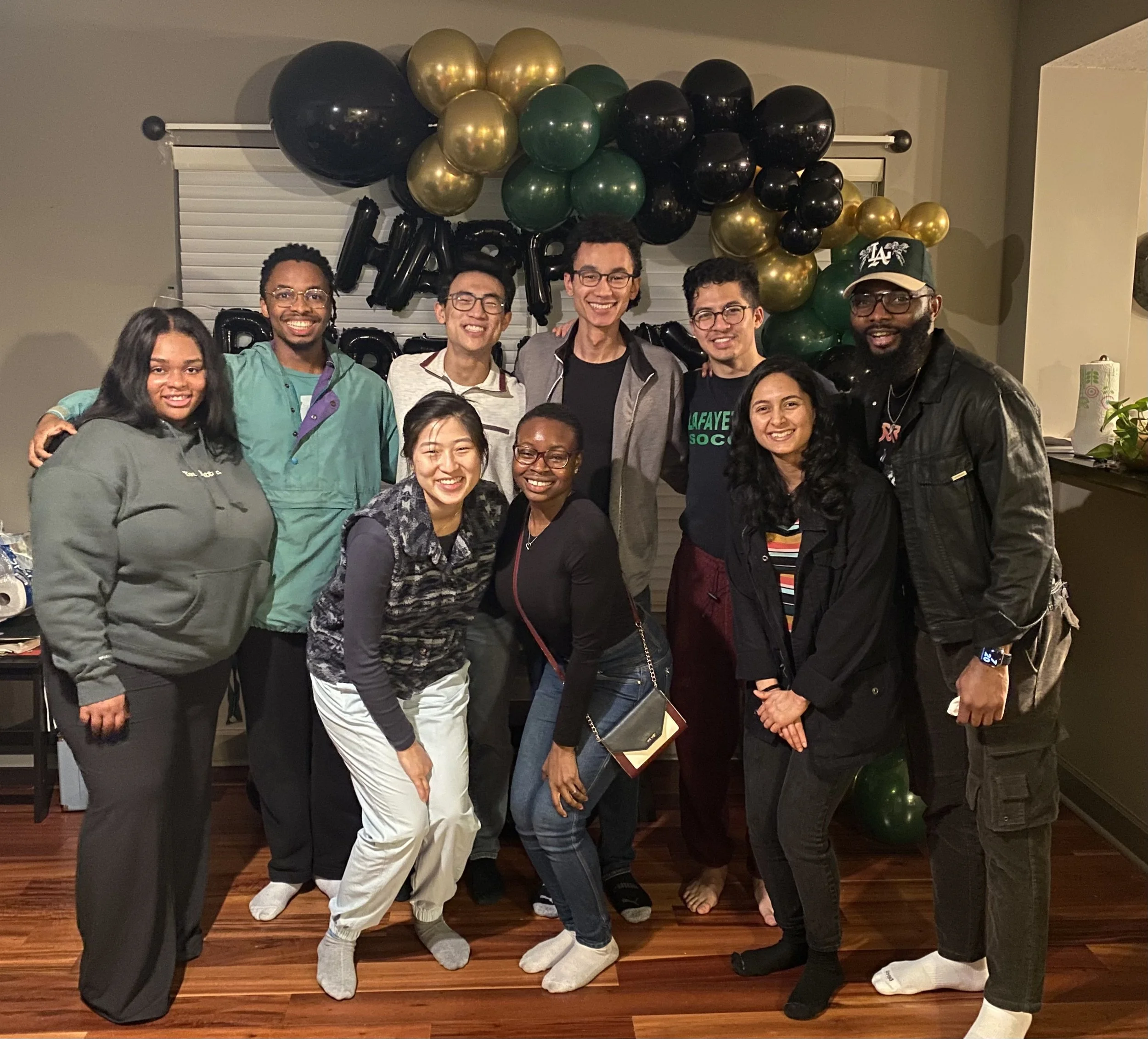 Group of Mope and Robert's friends smiling and posing for a photo indoors with black, gold, and green balloons in the background.