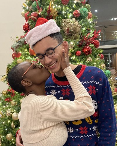 Mope and Robert, with Mope kissing Robert on the cheek, during a Christmas celebration. They are standing in front of a decorated Christmas tree with red and gold ornaments and lights.