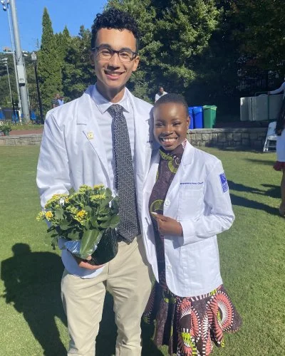 Mope and Robert in white coats standing outdoors on grass, smiling, with trees and blue sky in the background.