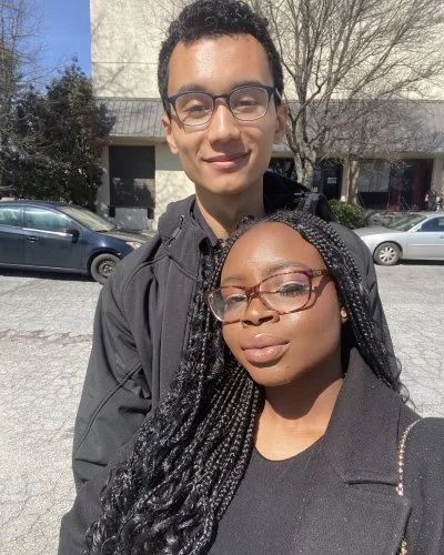 Robert and Mope posing outdoors for a selfie during the daytime, with parked cars and trees in the background.