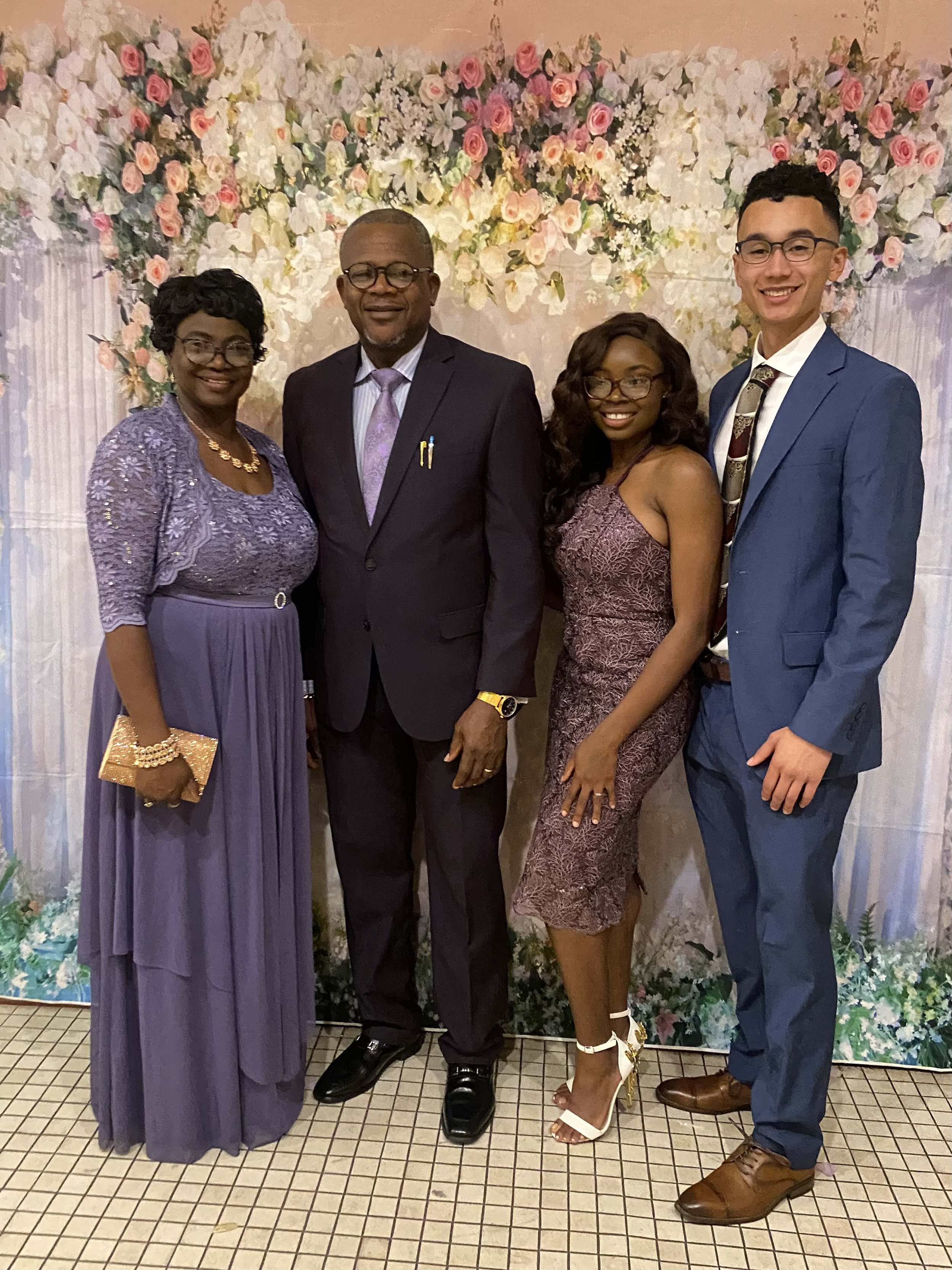 Mope and Robert with Mope's parents are dressed in formal attire posing for a photo in front of a floral backdrop.