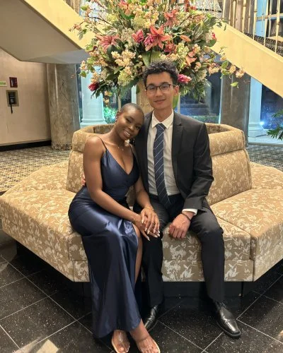 Robert and Mope in formal attire sitting on a floral-patterned sofa in a hotel lobby with a large floral arrangement in the background.
