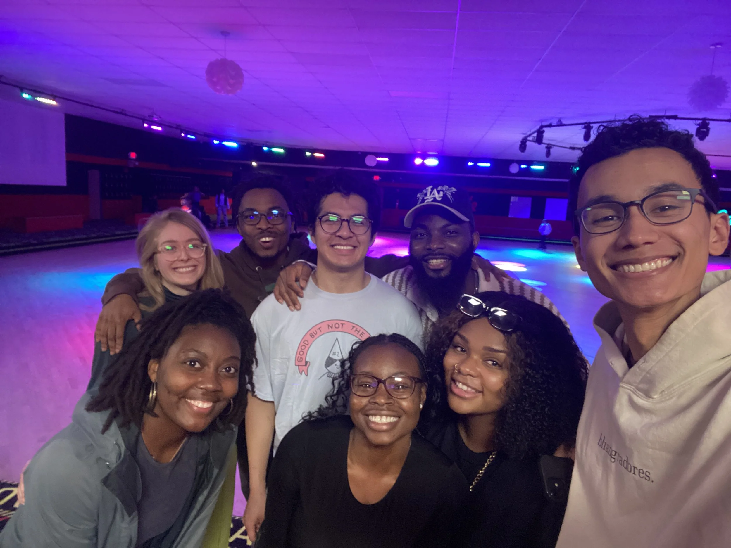 Group of Robert and Mope's friends, smiling and posing together at a roller skating rink with colorful lights and a spacious skating area in the background.