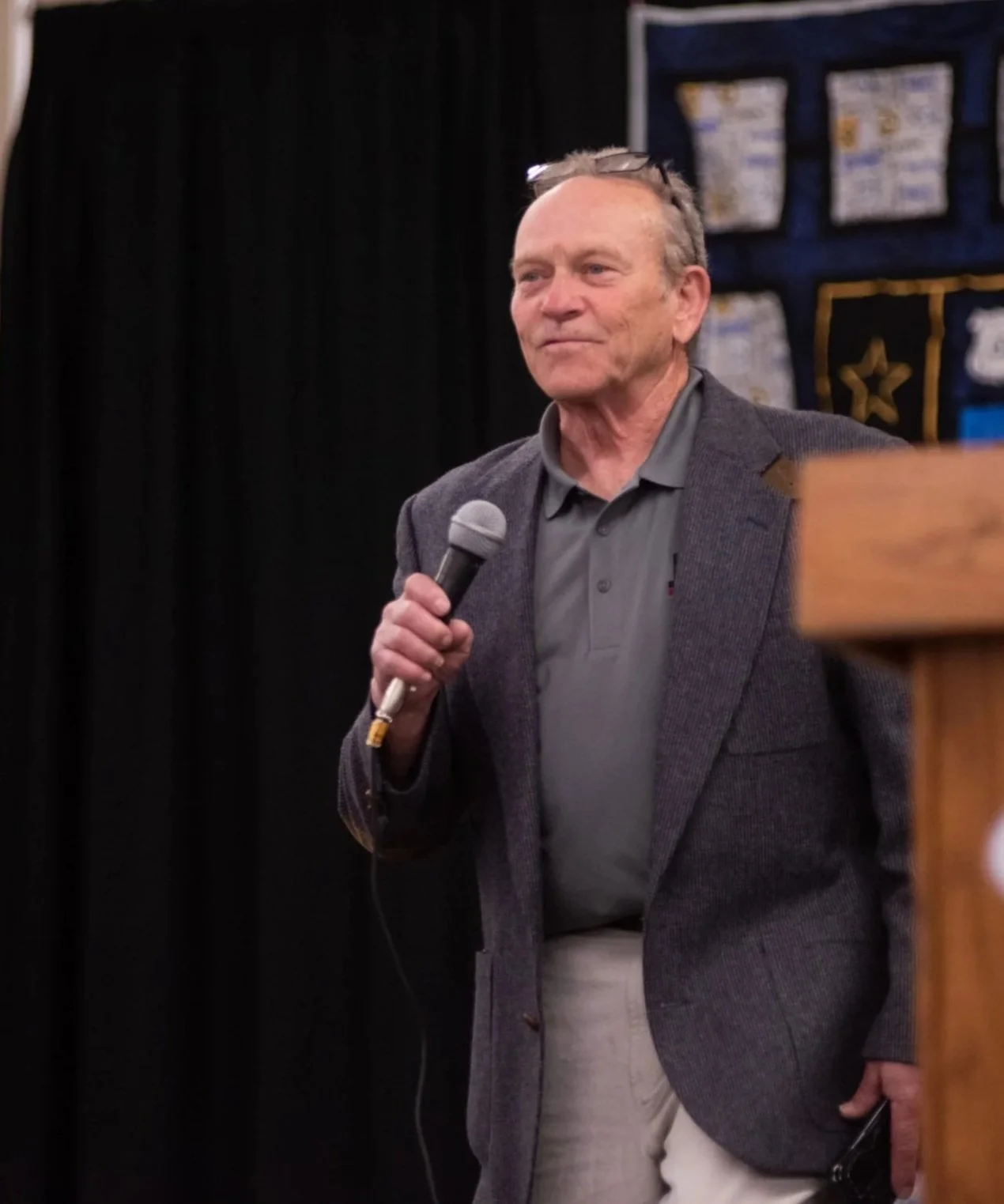 An elderly man in a gray blazer and gray polo shirt holding a microphone. He appears to be speaking at an event with a black curtain and display board in the background.