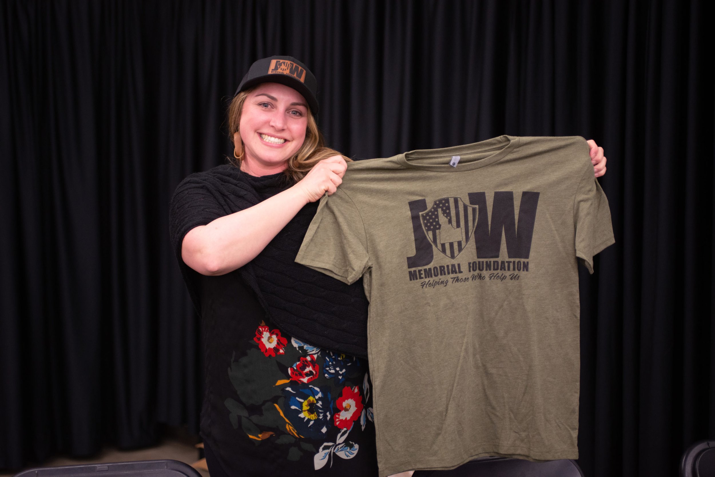 A woman smiling and holding up a khaki T-shirt with 'JW Memorial Foundation Helping Those Who Help Us' printed on it, standing in front of a black curtain.