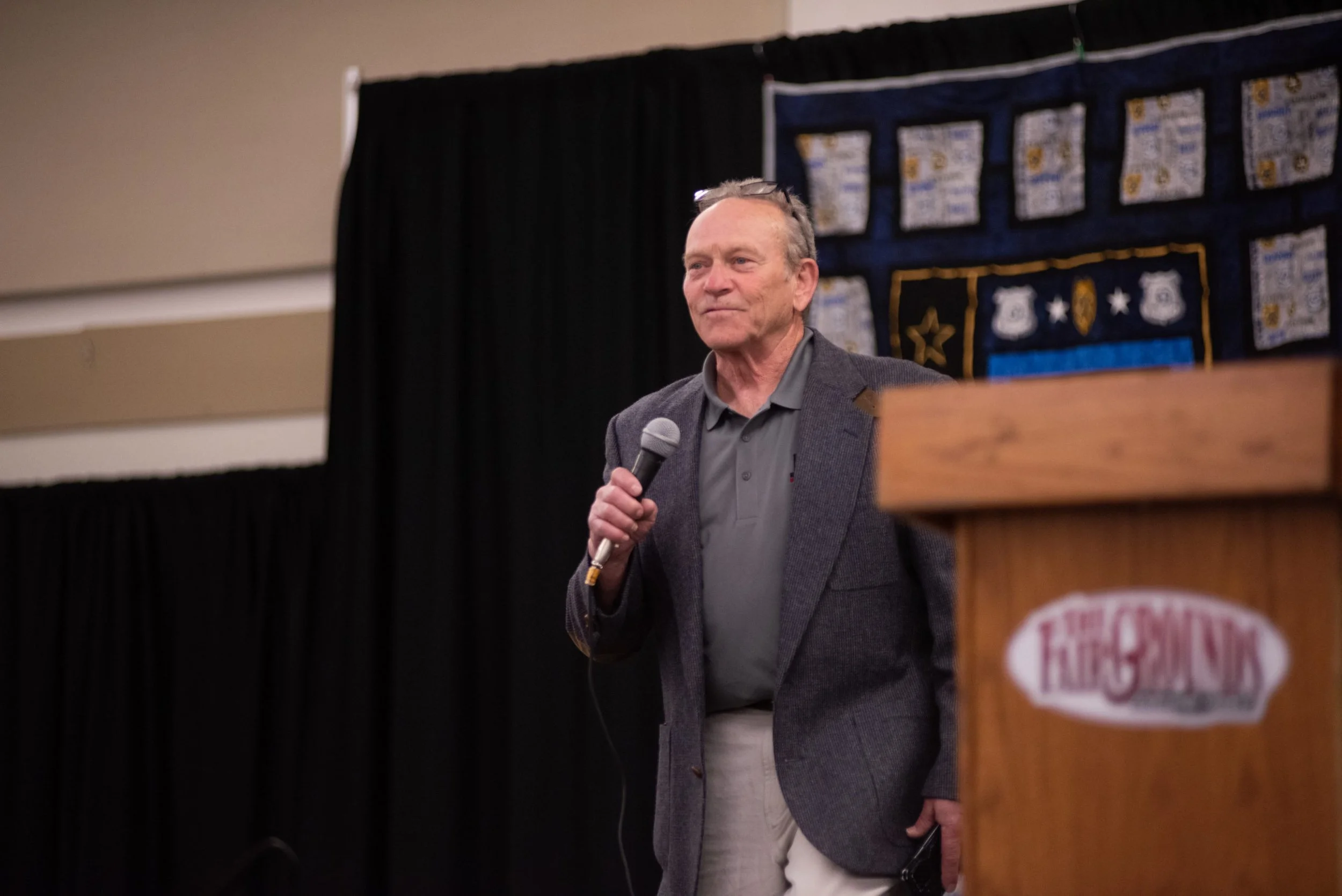 An older man holding a microphone stands on stage during a presentation or speech.