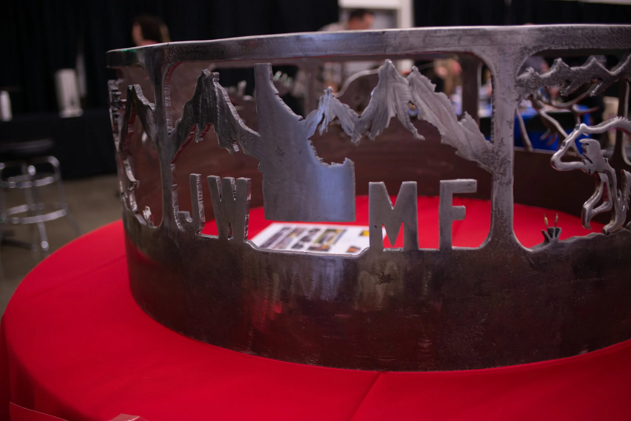 Metal wall art with cutouts of trees, mountains, and 'JWMF', placed on a red tablecloth, with a blurred background of a room with people.