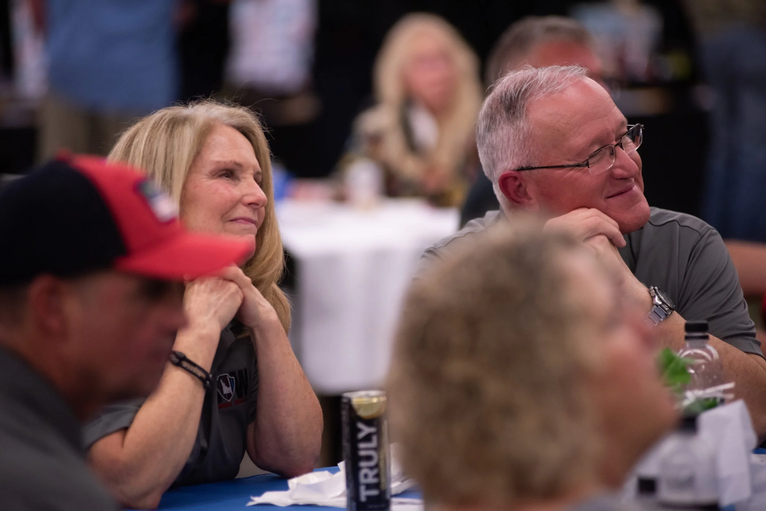 People sitting at a table in an indoor setting, some smiling and others with neutral expressions, with drinks and papers on the table.