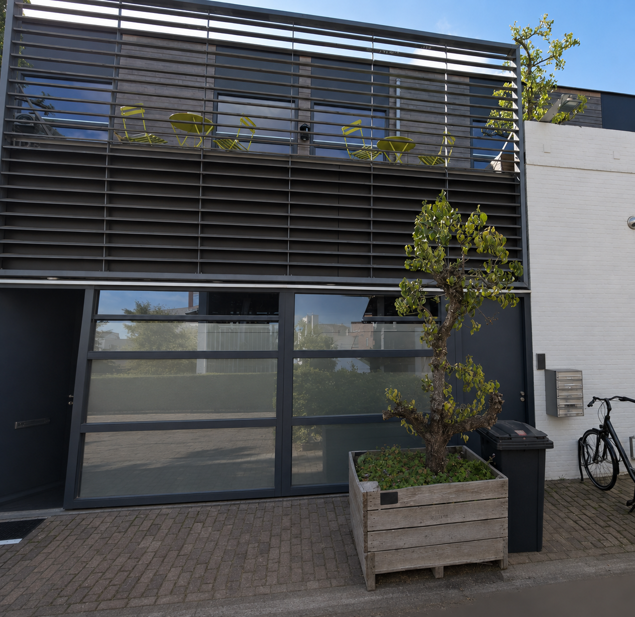 Modern gebouw met grote ramen en een balkon met gele tuinmeubels, een kleine boom in een houten planter en een fiets.