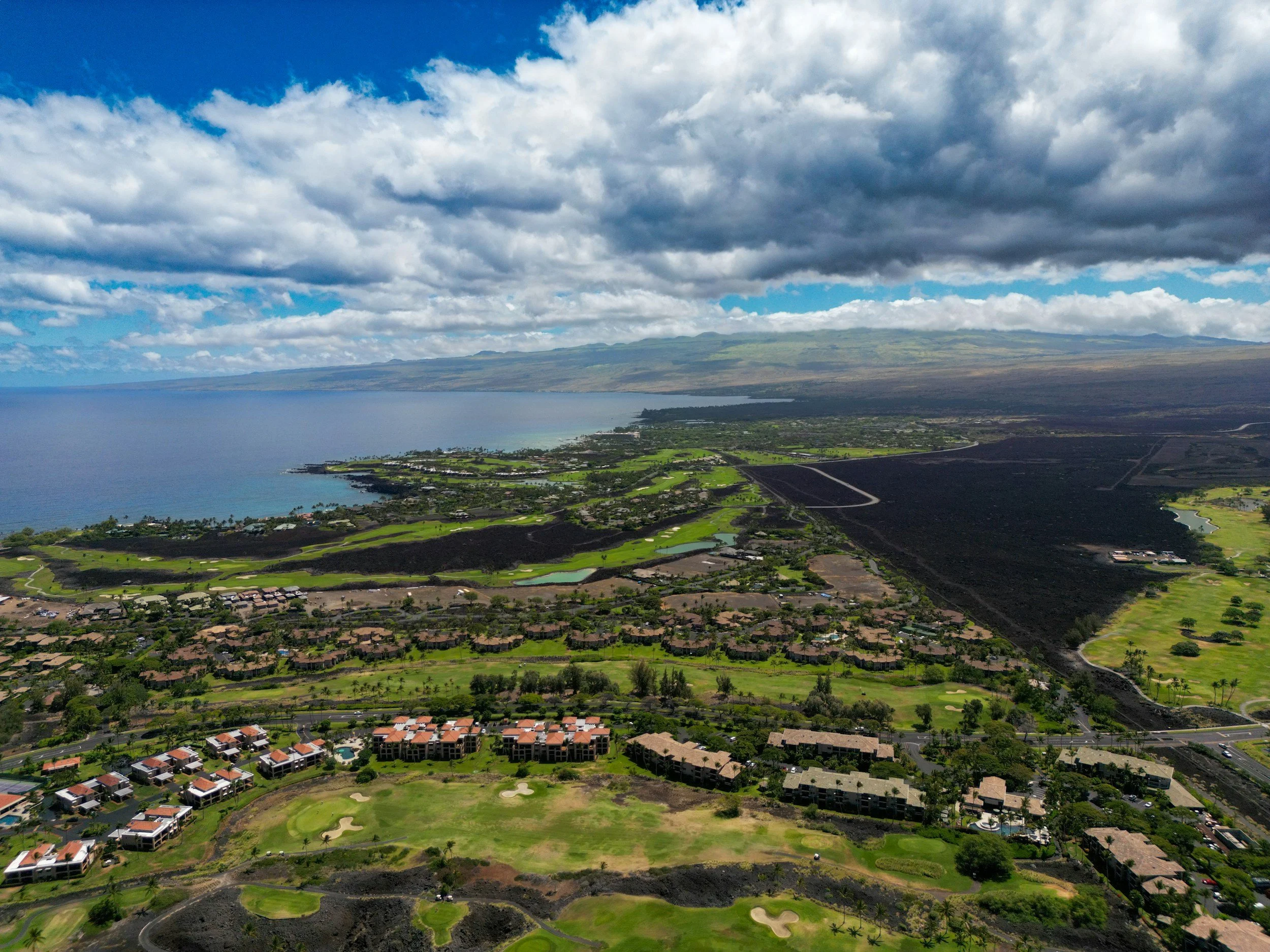 Aerial view of a coastal landscape with houses, golf courses, green fields, black volcanic lava, and the ocean under a partly cloudy sky.