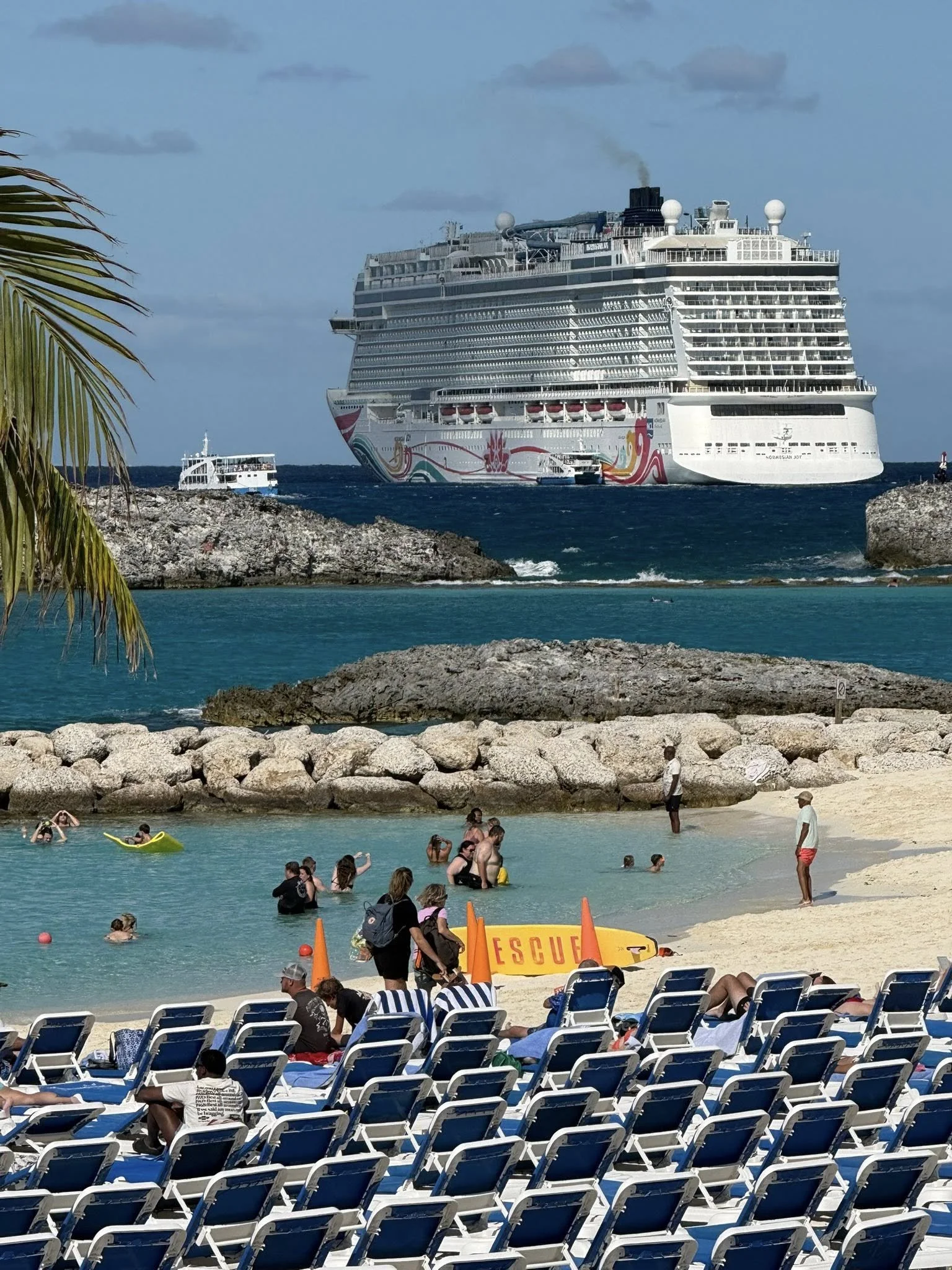 People relaxing on lounge chairs and swimming in a beach with a large cruise ship in the background.